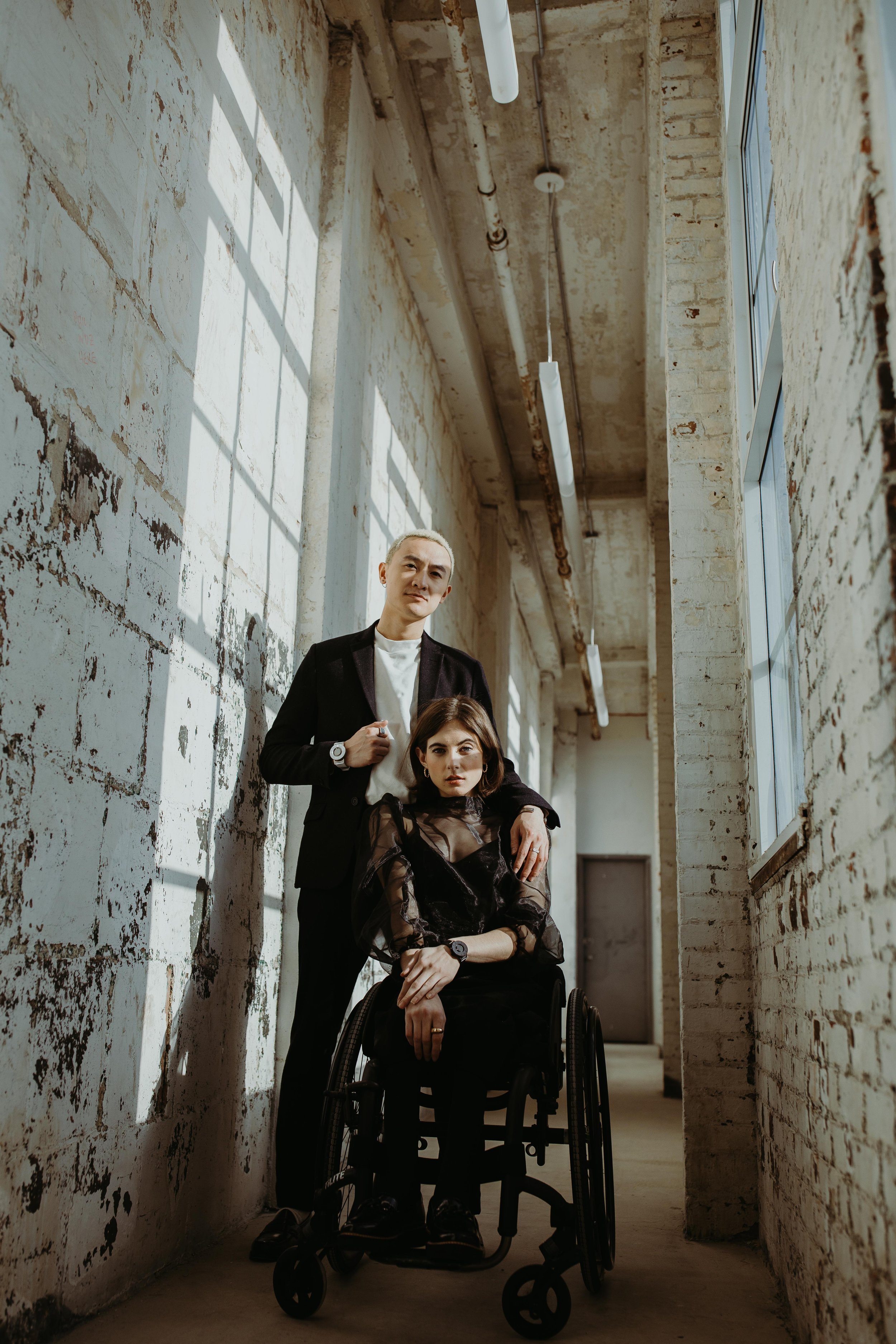 Two people in an industrial hallway, one standing and the other seated in a wheelchair, wearing stylish clothing.