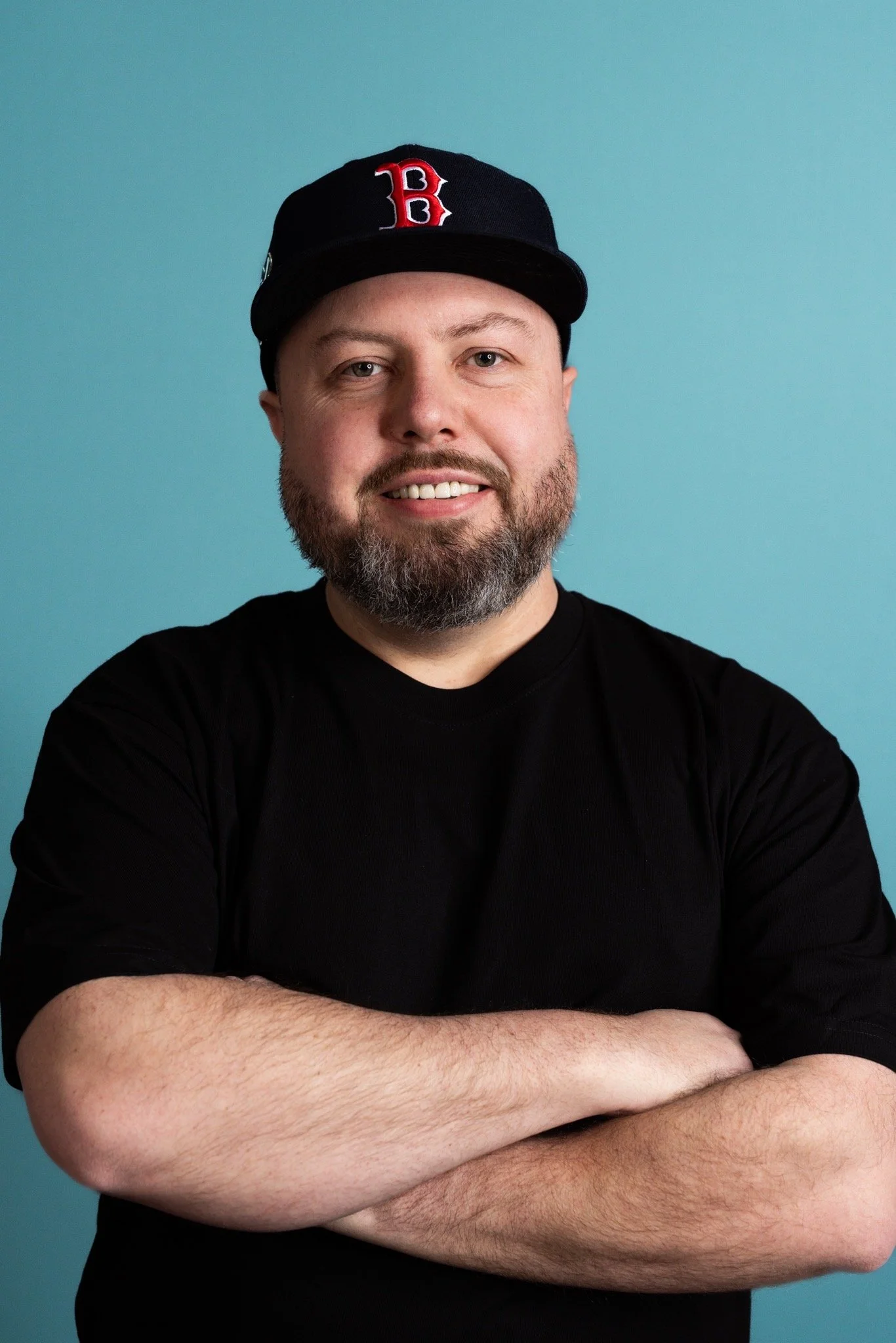 Person wearing a black shirt and a Boston Red Sox cap, smiling with arms crossed, standing against a blue background.