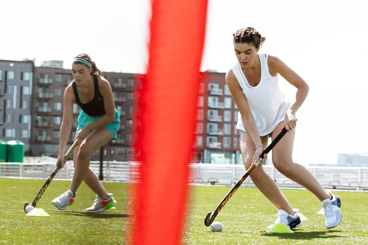 Two female field hockey players in action on a grass field, with buildings in the background.