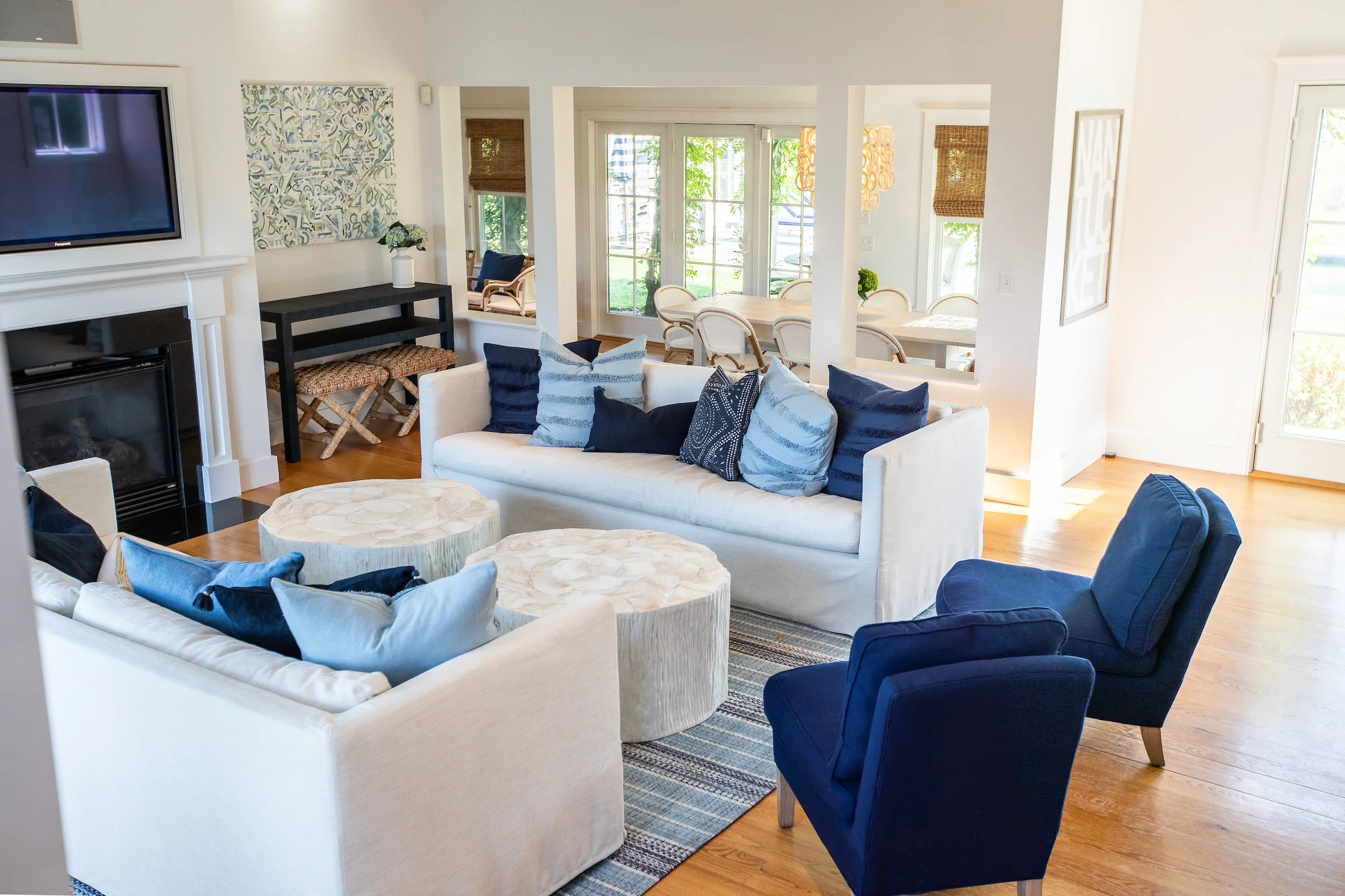 Coastal-style living room with white and blue furniture, including sofas and armchairs, a fireplace, and a TV. Light-colored wooden floor, decorative pillows, and a dining area visible in the background with rattan chairs and a table.