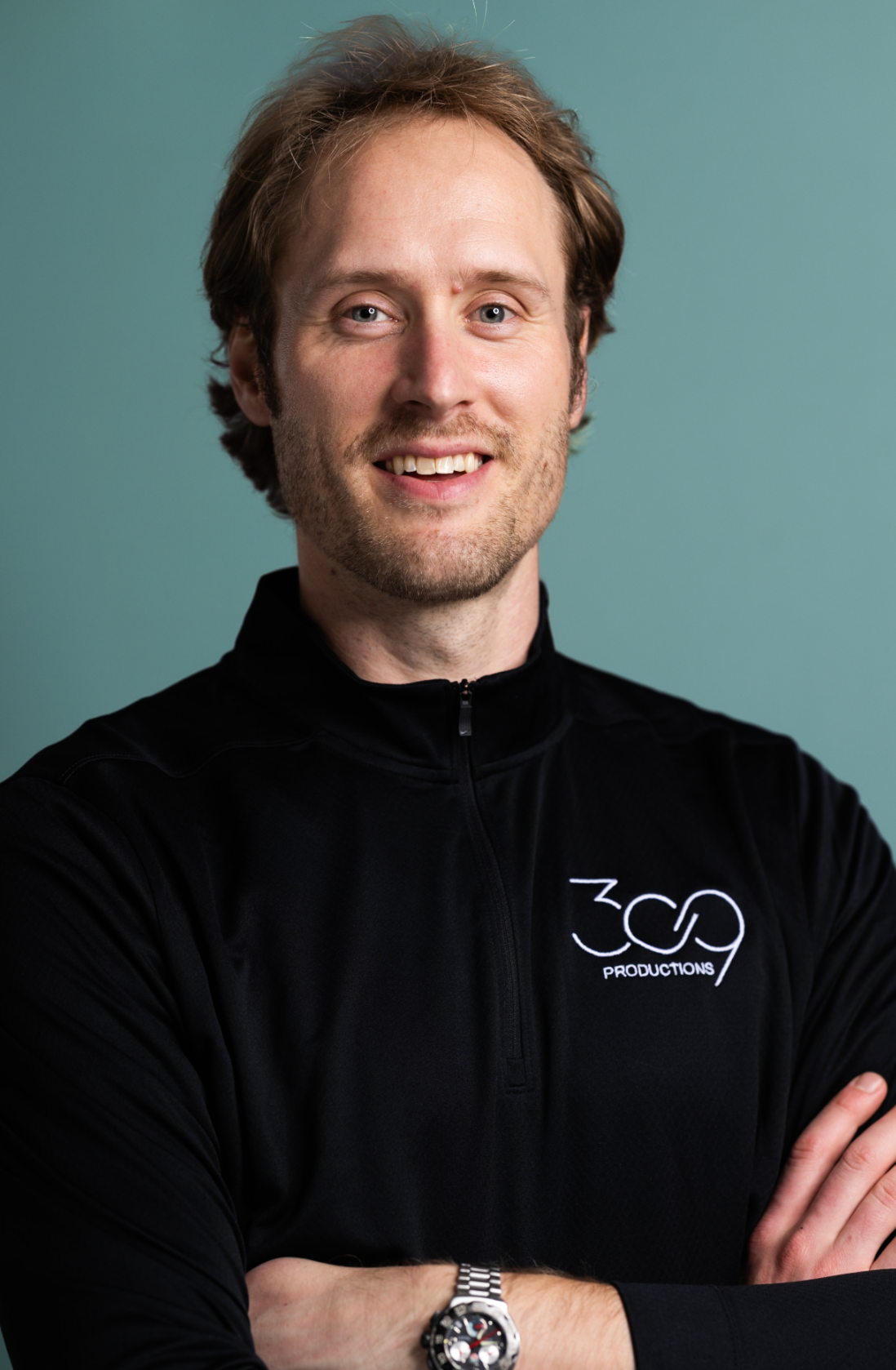 Smiling man in a black shirt with "369 Productions" logo, arms crossed, against a teal background.
