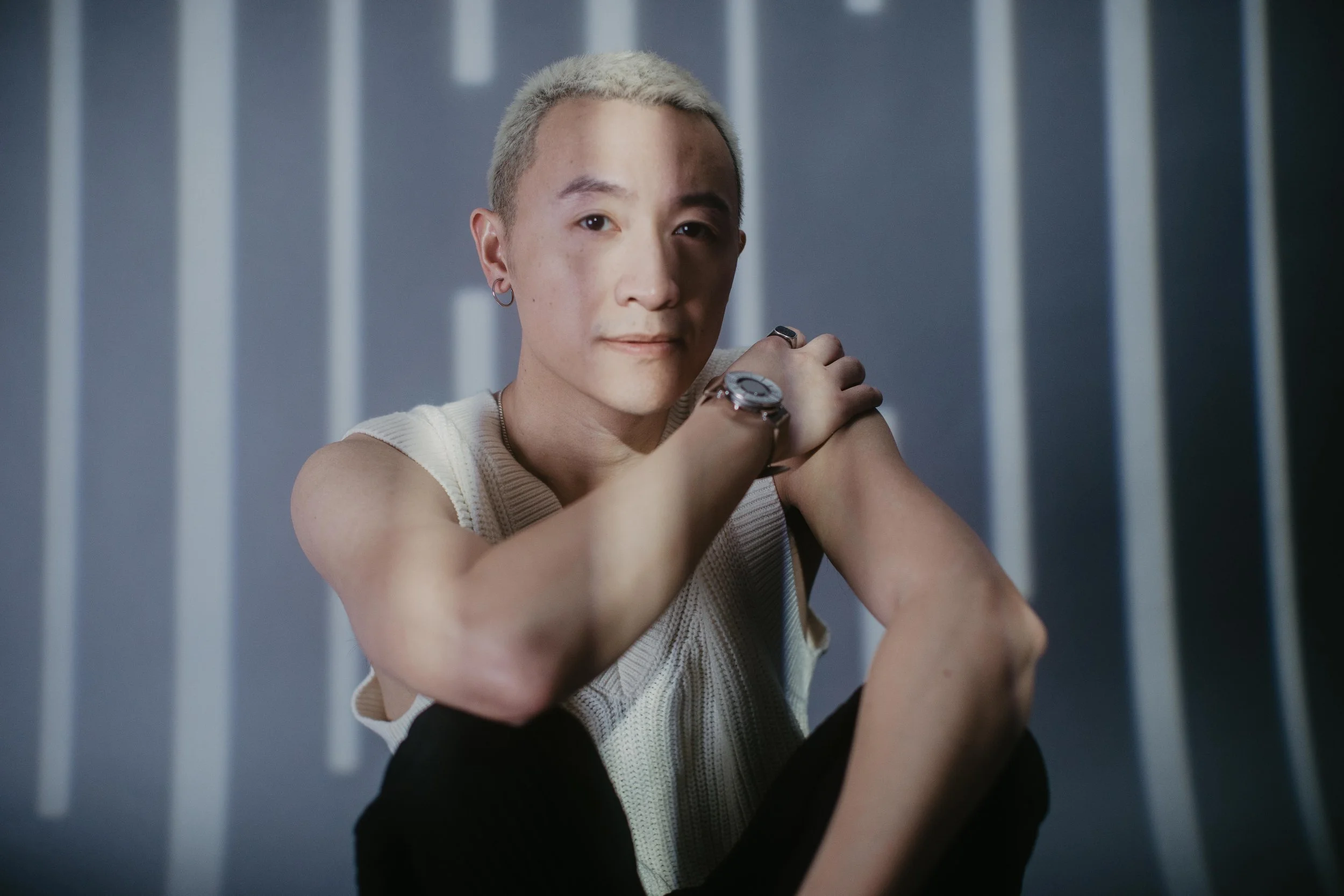 Person wearing a sleeveless top and watch, seated with one arm crossed, in front of a striped background.