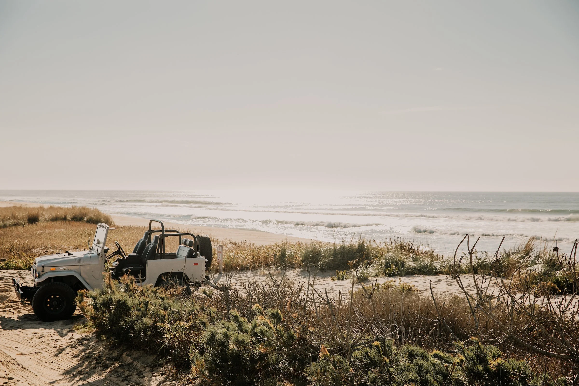 Open jeep parked on a sandy beach by the ocean, with waves in the background and vegetation around.