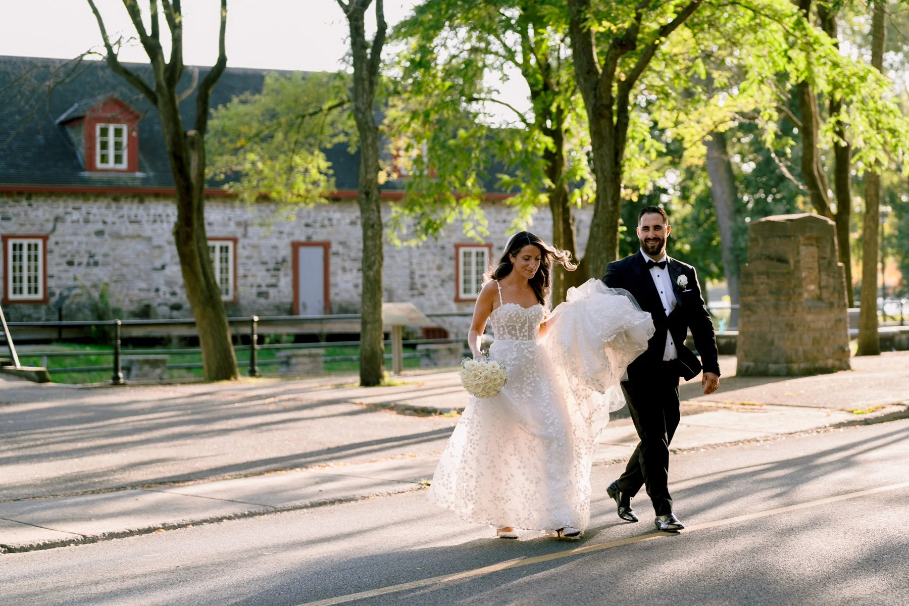 Newlywed couple walking together outdoors during their couple photo session after their montreal wedding ceremony, with the bride carrying her bouquet and dress backlit by sunlight.