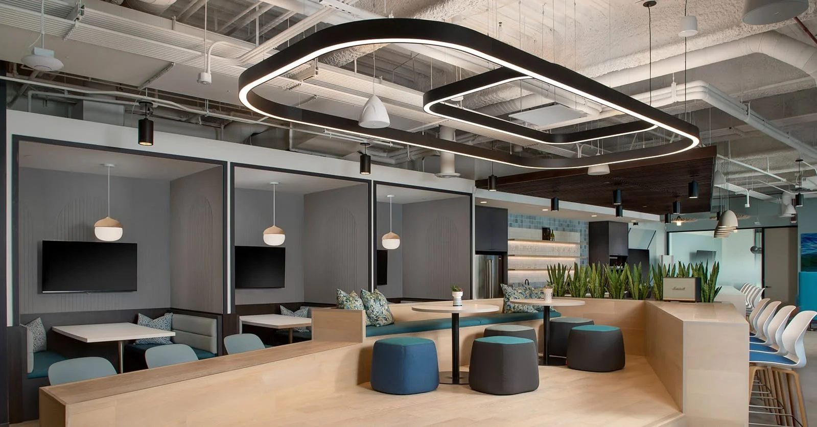 Modern office lounge area with tables, chairs, and hanging pendant lights. There are sofas with cushions, potted plants, and a decorative ceiling light fixture. The space features bright, contemporary design elements and an open layout.
