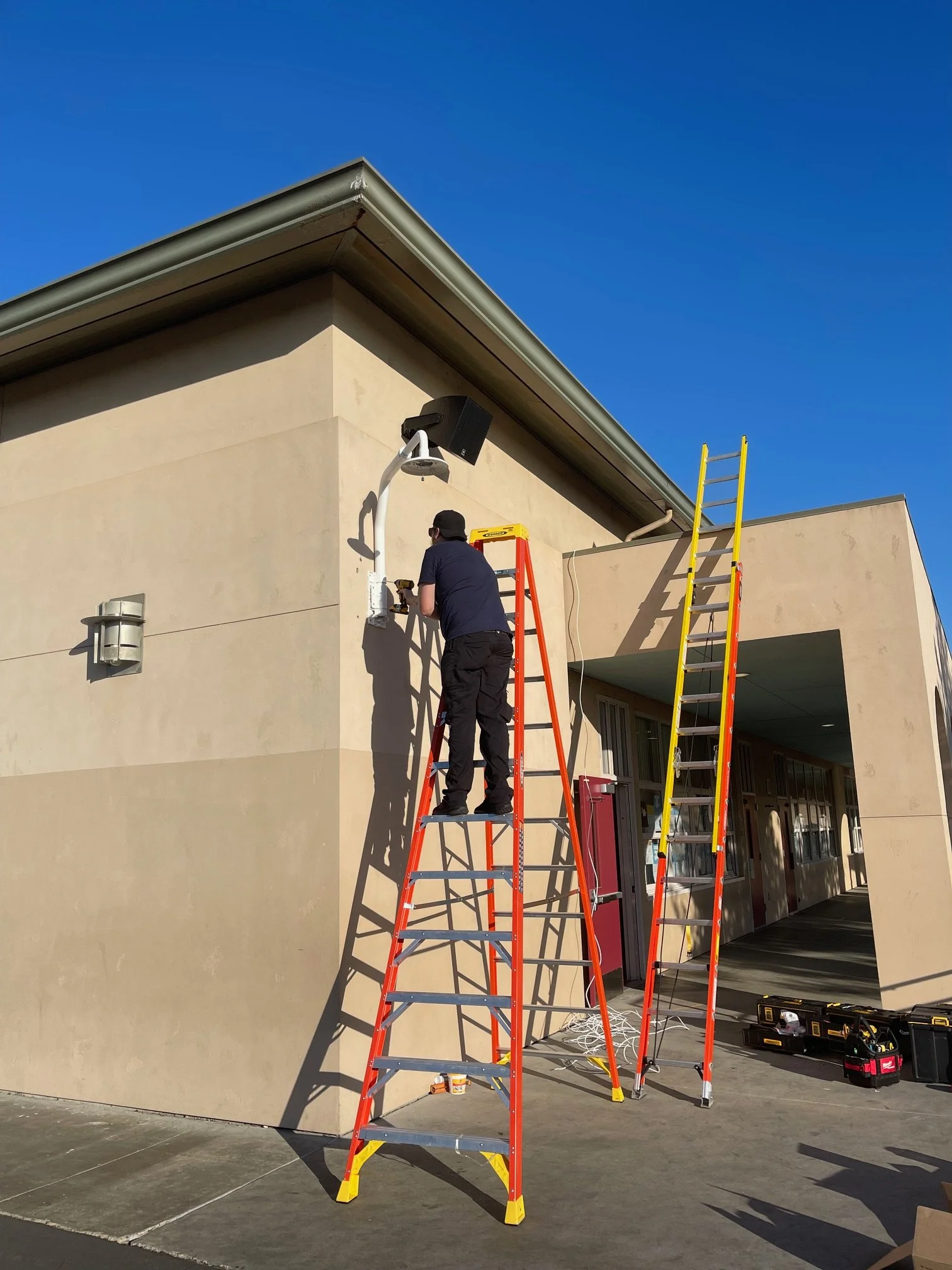 A person working on an outdoor wall, using a ladder and power tools, with a second ladder nearby and equipment on the ground, under clear blue sky.