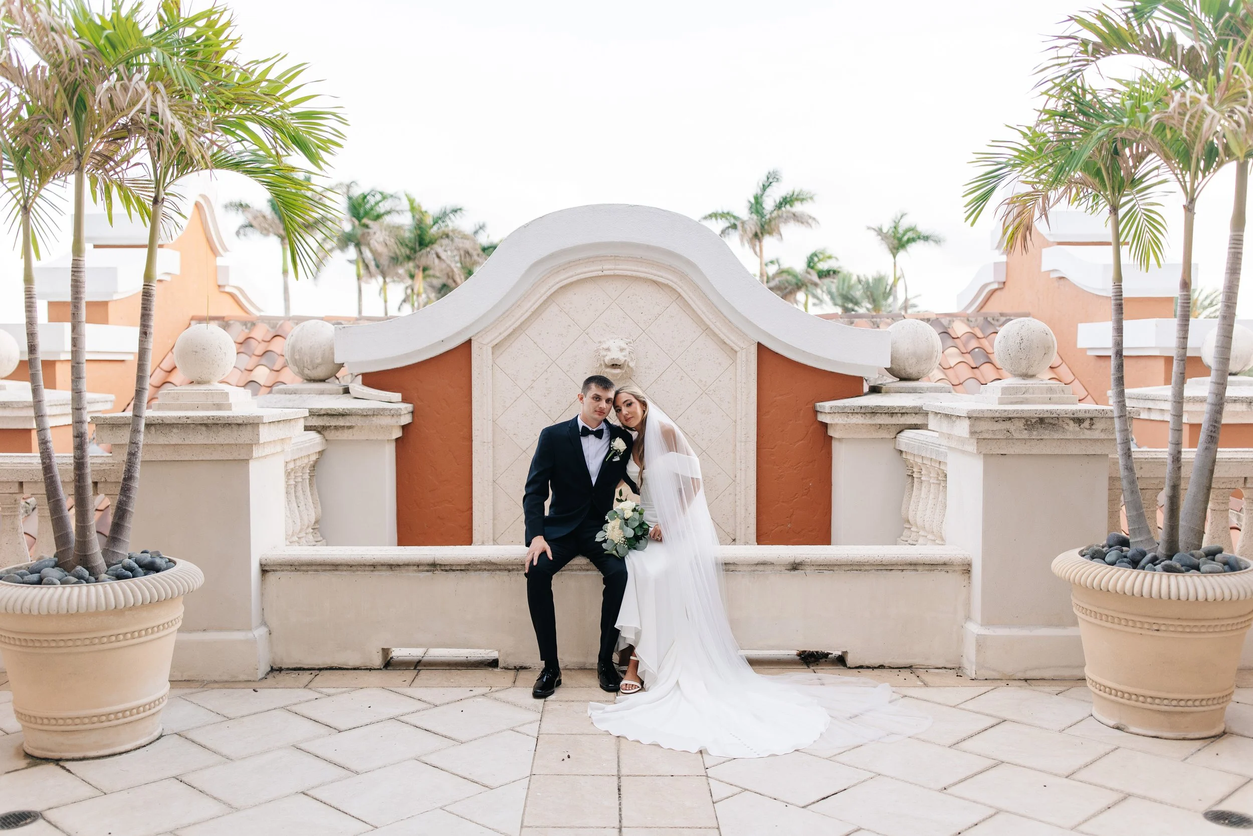 Bride and groom sitting together on a palm-lined terrace at their Palm Beach wedding venue 