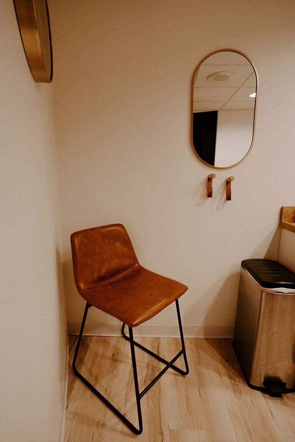 A cozy corner with a brown leather chair, a small mirror, and a trash bin in a room with wooden flooring  at Salt + Light Salon and Spa in Cincinnati, OH