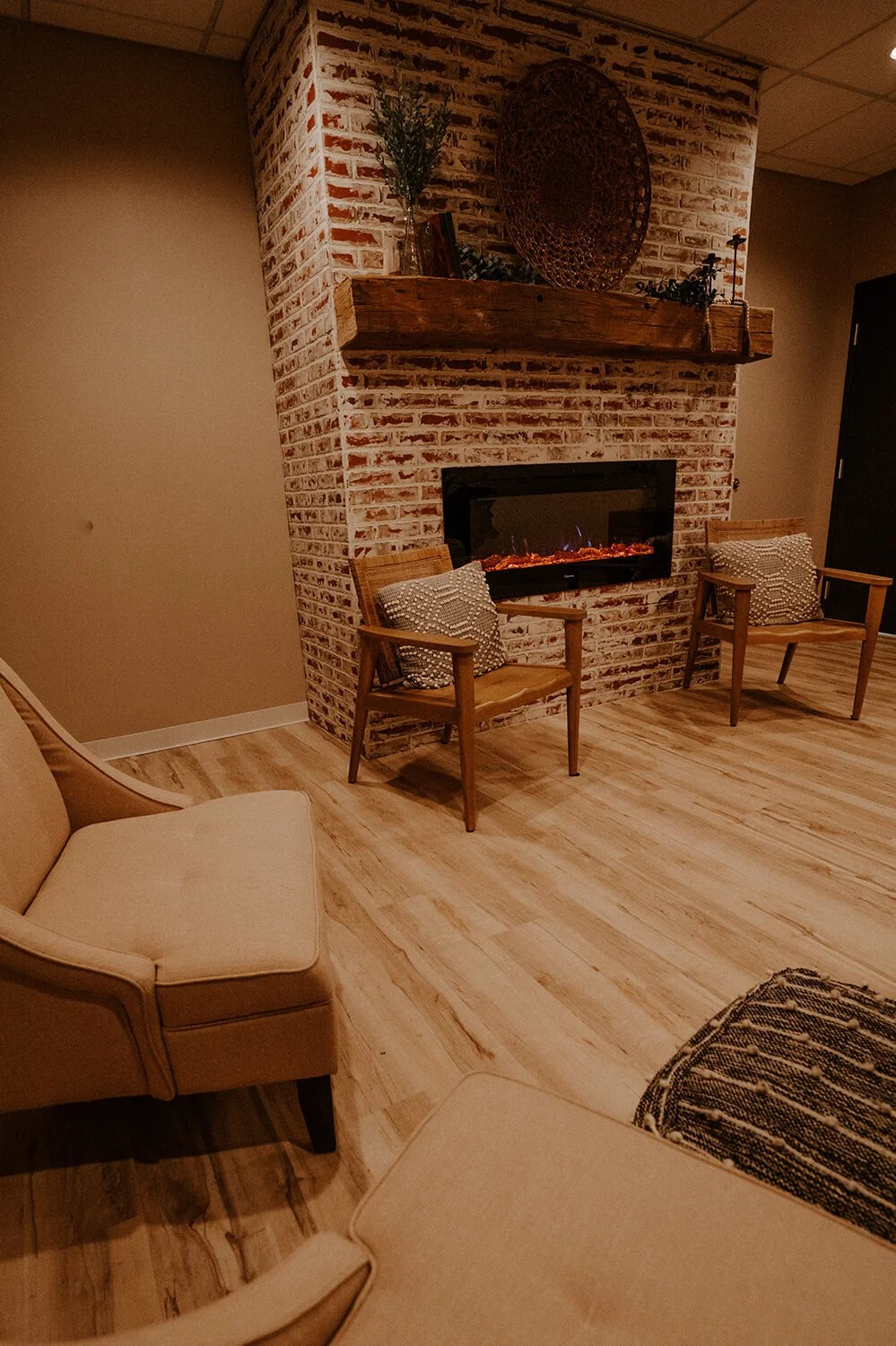 Waiting room with brick fireplace, wooden mantel, two wooden chairs with cushions, beige sofa, and a patterned rug on wood floor  at Salt + Light Salon and Spa in Cincinnati, OH
