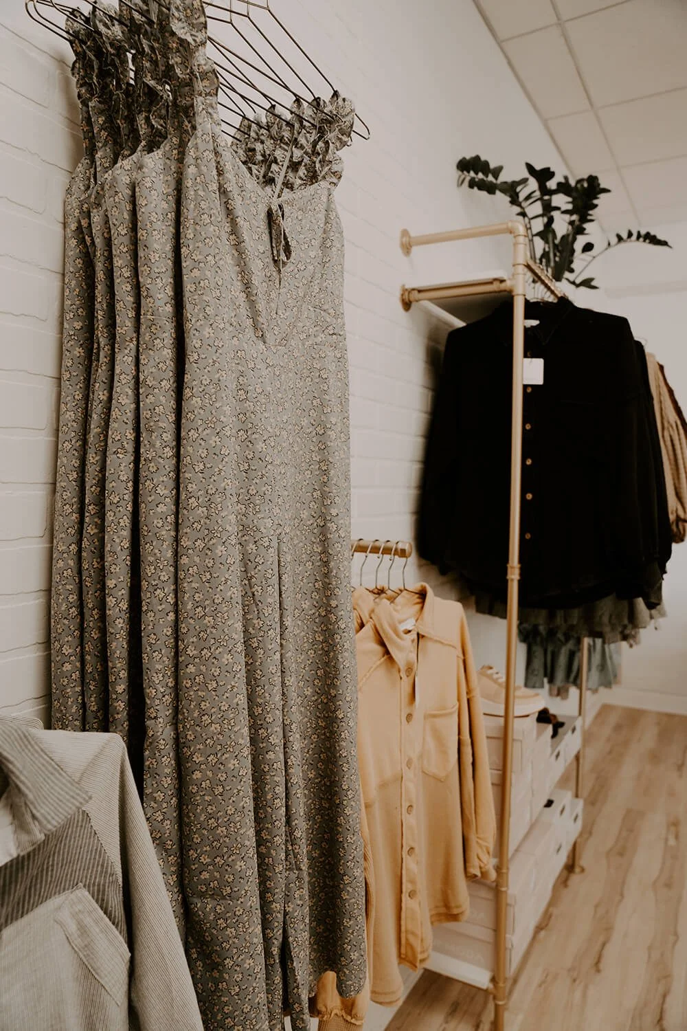 A floral dress hanging on a wire hanger, a yellow jacket on a wooden rack, and black and beige clothing on a clothing rack display against a white wall at the Shop Salt boutique.