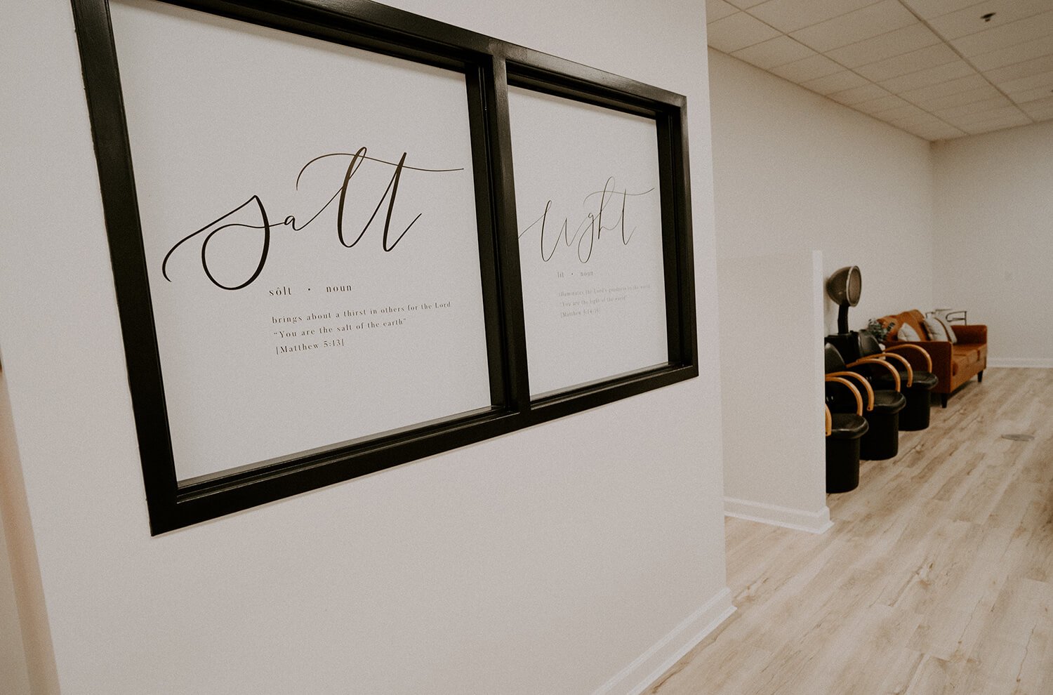 Decorative framed spelling words 'salt' and 'light' with definitions and Bible verse shown on a white wall in a room with wooden flooring and seating area  at Salt + Light Salon and Spa in Cincinnati, OH