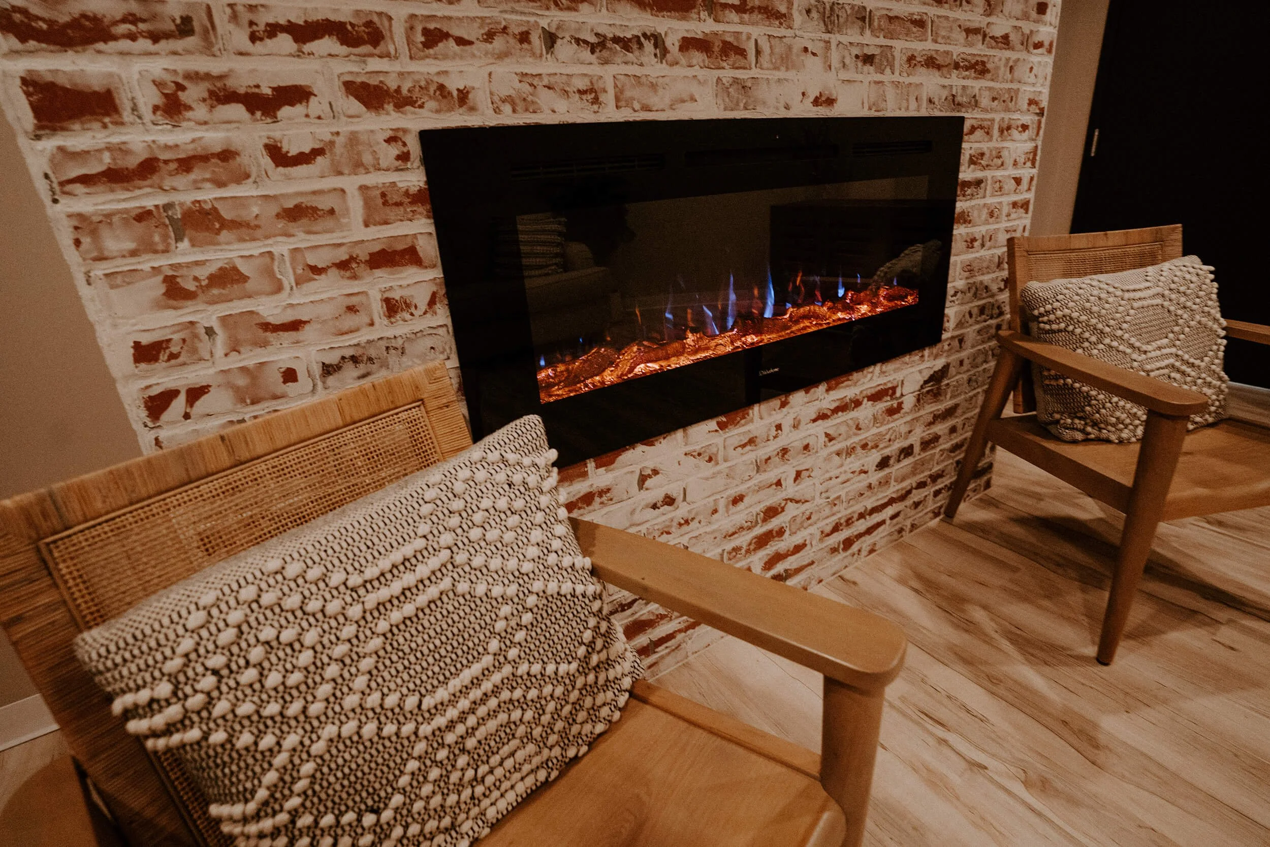 Waiting room with two wooden chairs with woven seat cushions, a brick wall with a modern electric fireplace in the center, and wood flooring  at Salt + Light Salon and Spa in Cincinnati, OH