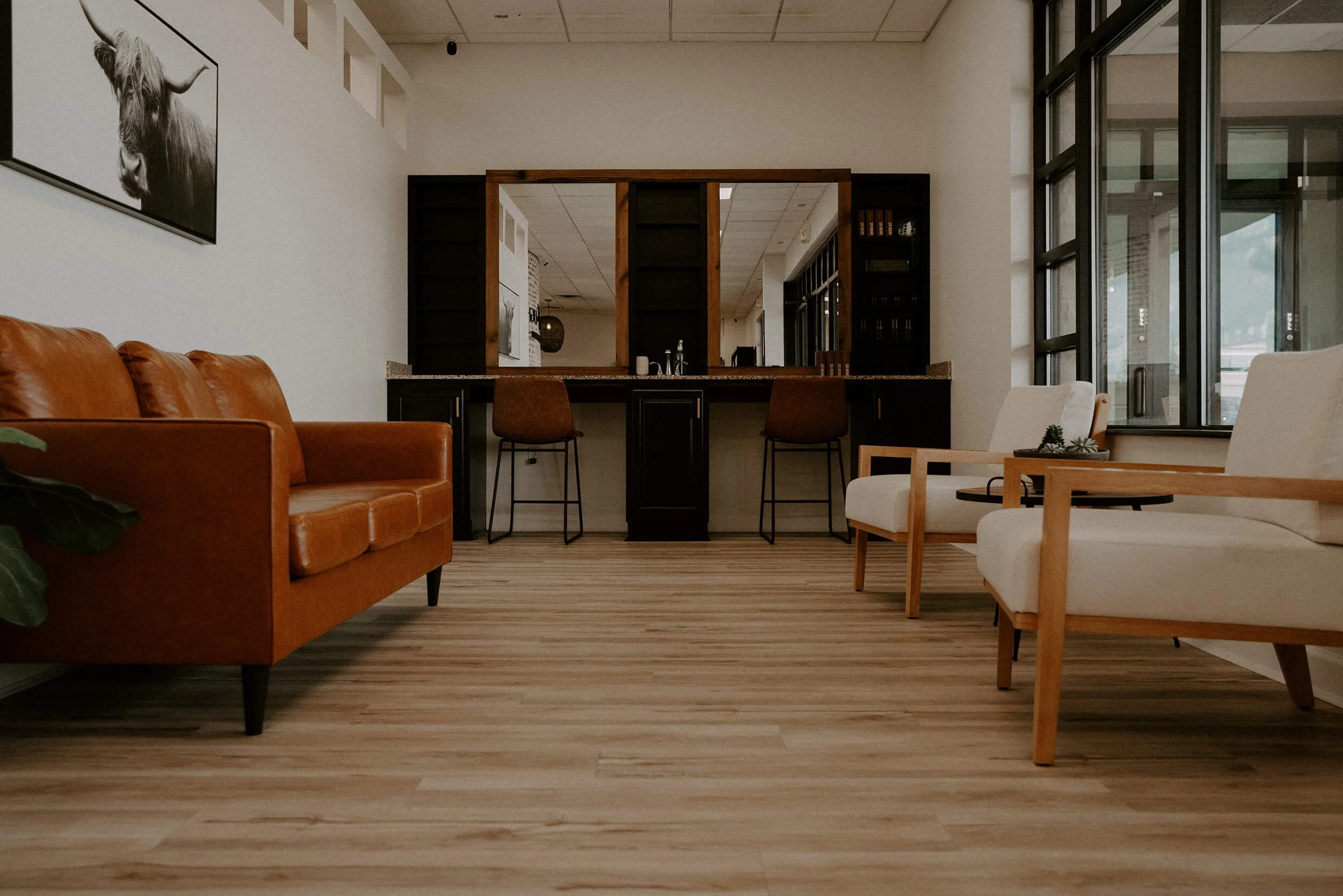 Modern waiting room with wooden flooring, leather sofa, white armchairs, and large windows at Salt + Light Salon and Spa in Cincinnati, OH