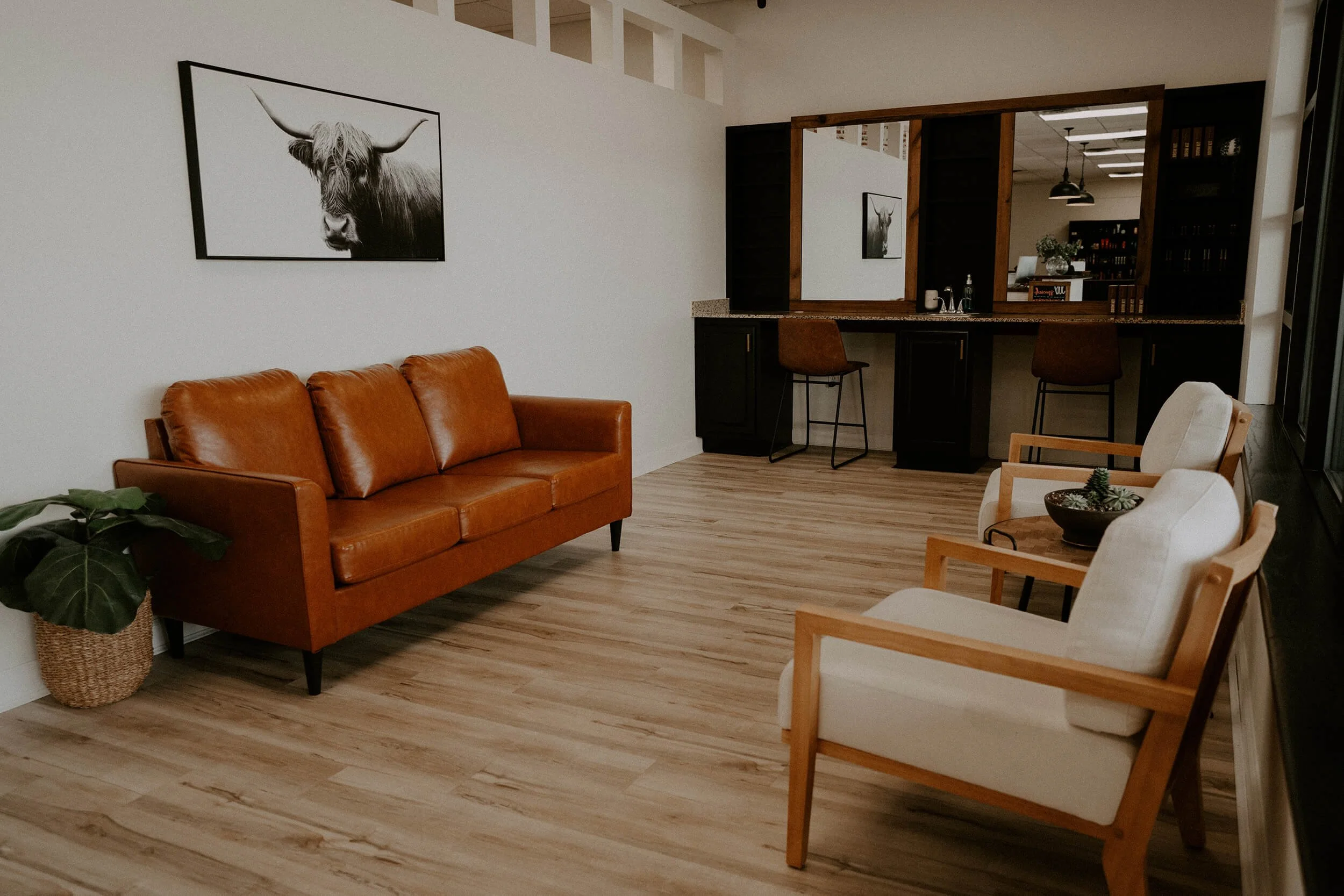 Modern waiting room with a brown leather sofa, two white armchairs, a potted plant, two brown chairs, and a mirror  at Salt + Light Salon and Spa in Cincinnati, OH