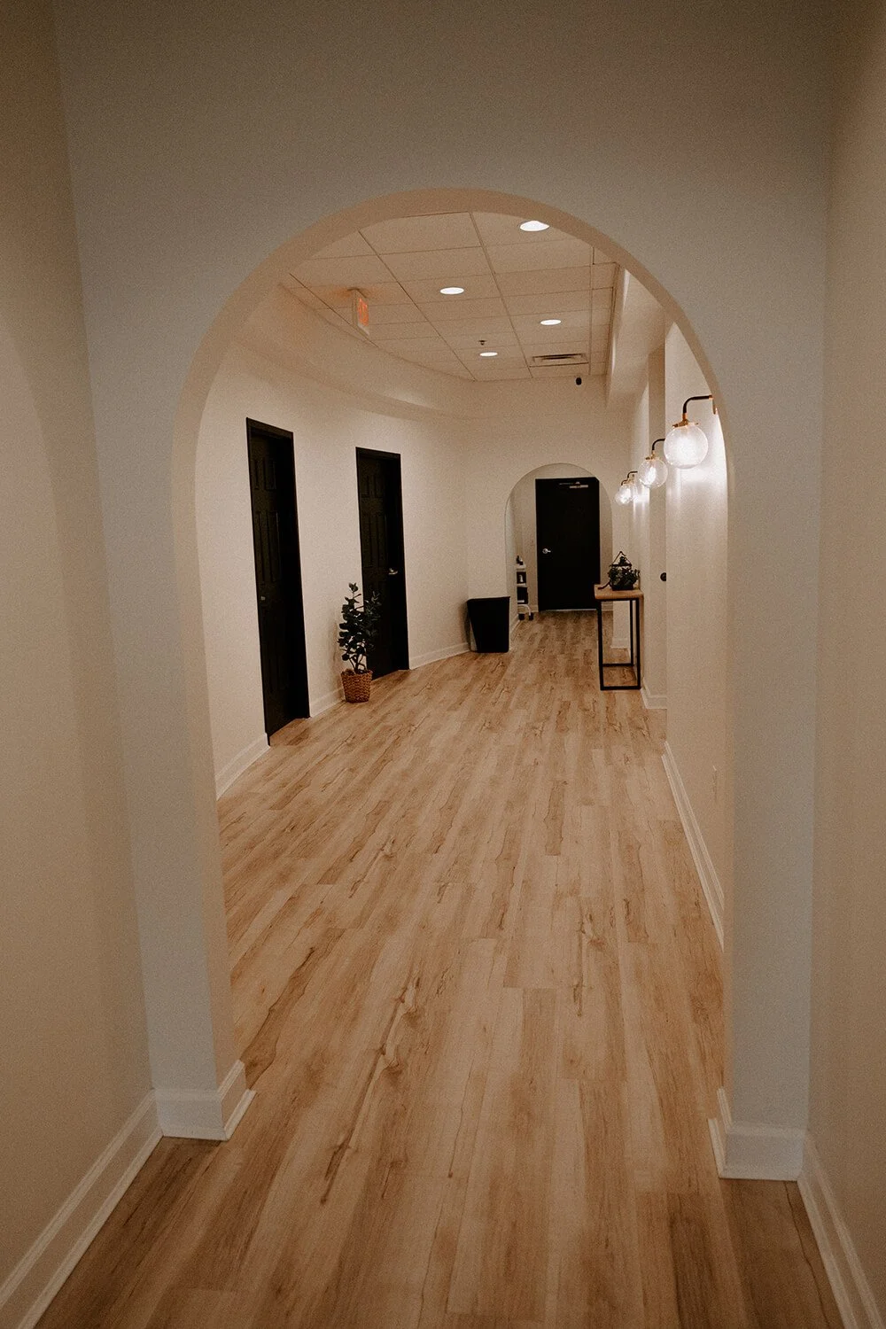 Hallway with wooden flooring, white walls, black doors, small potted plant, and black table with decorative items  at Salt + Light Salon and Spa in Cincinnati, OH