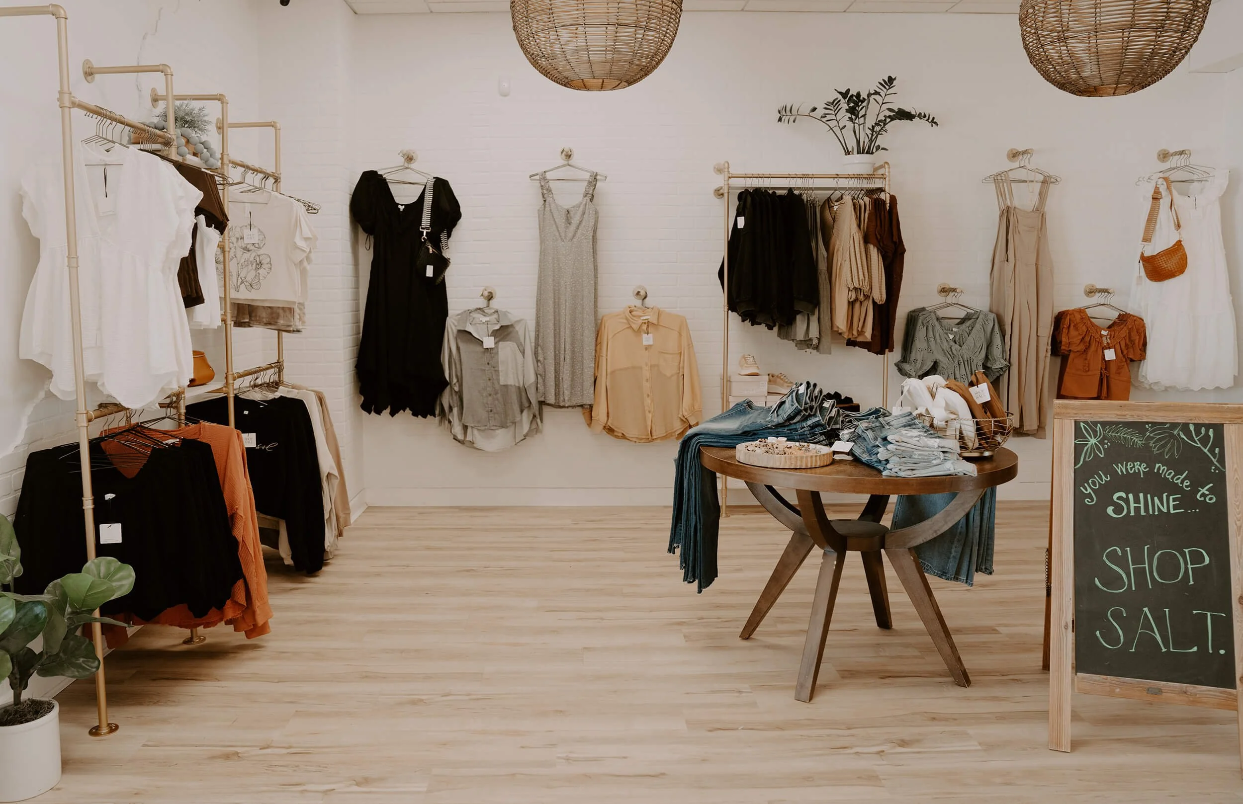 Clothing boutique with beige racks and shelves displaying various women's clothes like dresses, blouses, and pants. A round wooden table in the center holds folded denim and accessories. A chalkboard sign reads 'You were made to shine... Shop Salt.'