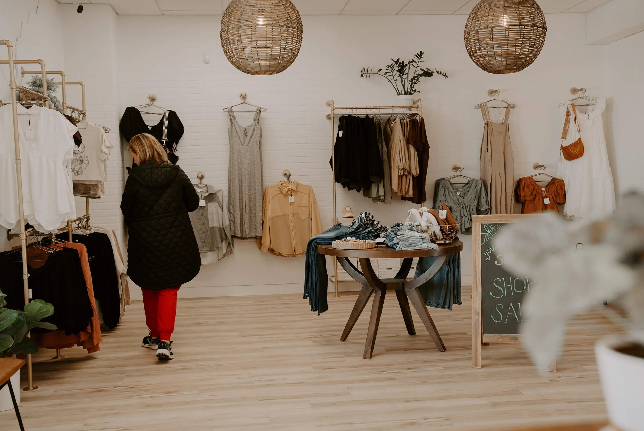 Interior of the Shop Salt boutique with various dresses, tops, and pants hanging on racks against white brick walls, a wooden table with folded clothes and accessories, a chalkboard sign advertising a sale, and a customer browsing.