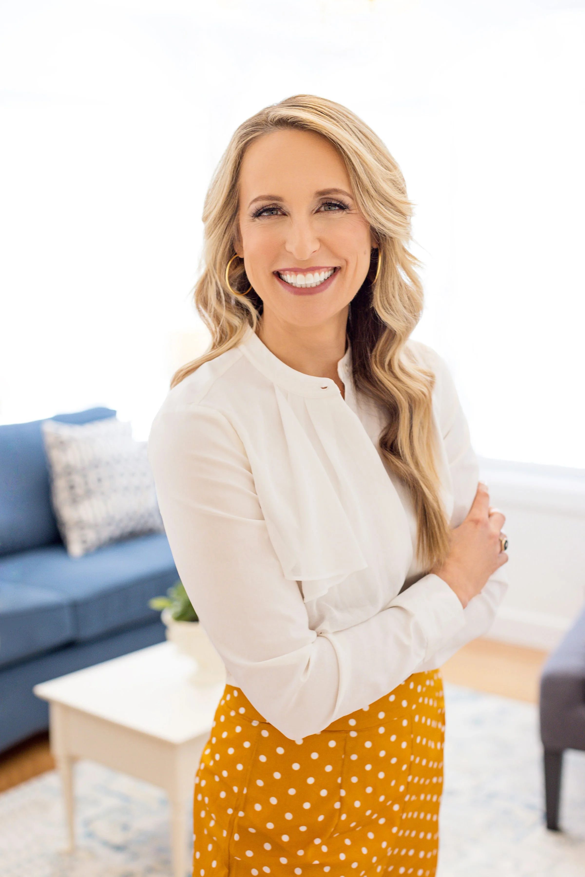 woman with long blond hair wearing a button down white shirt and yellow flowing pants with white polka dots, standing with arms crossed at the waist in the center of her office