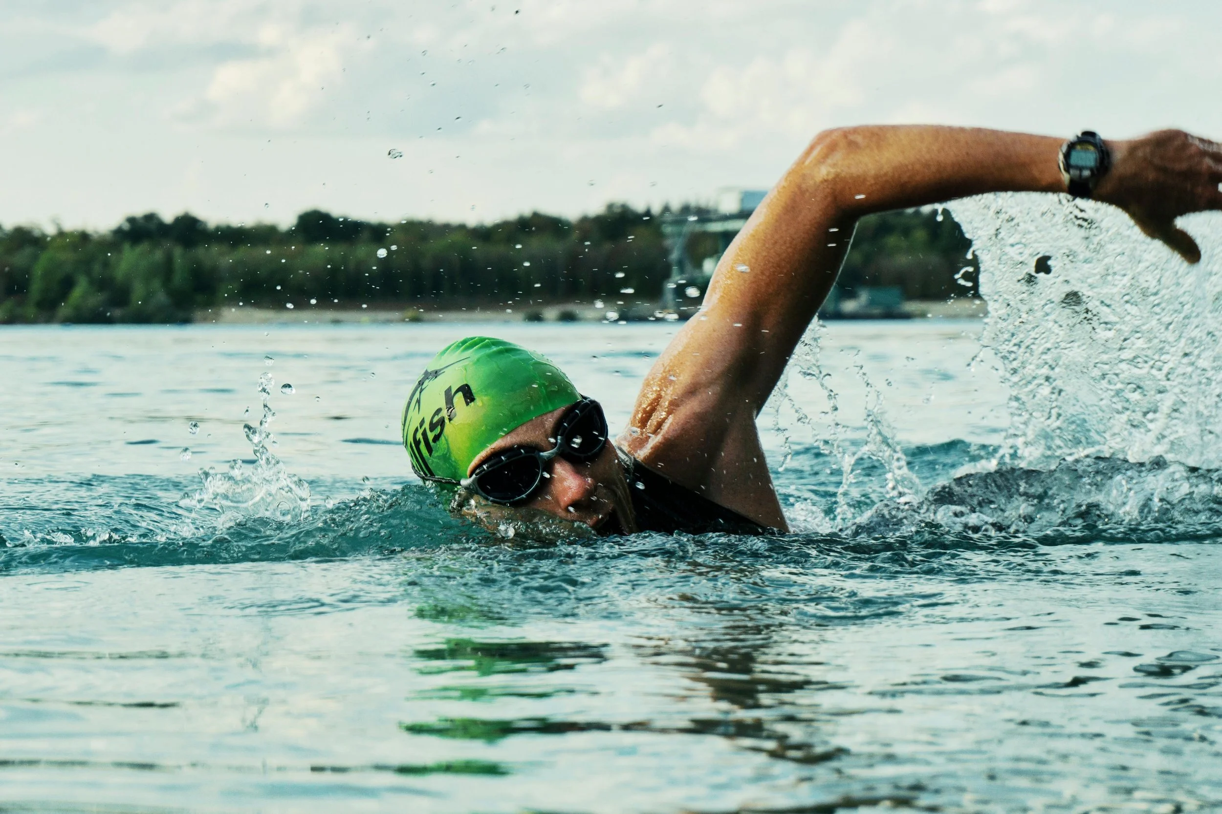 Athletic Man with a green swim cap and black googles swims mid freestyle stroke through an open lake with trees in the distant shore