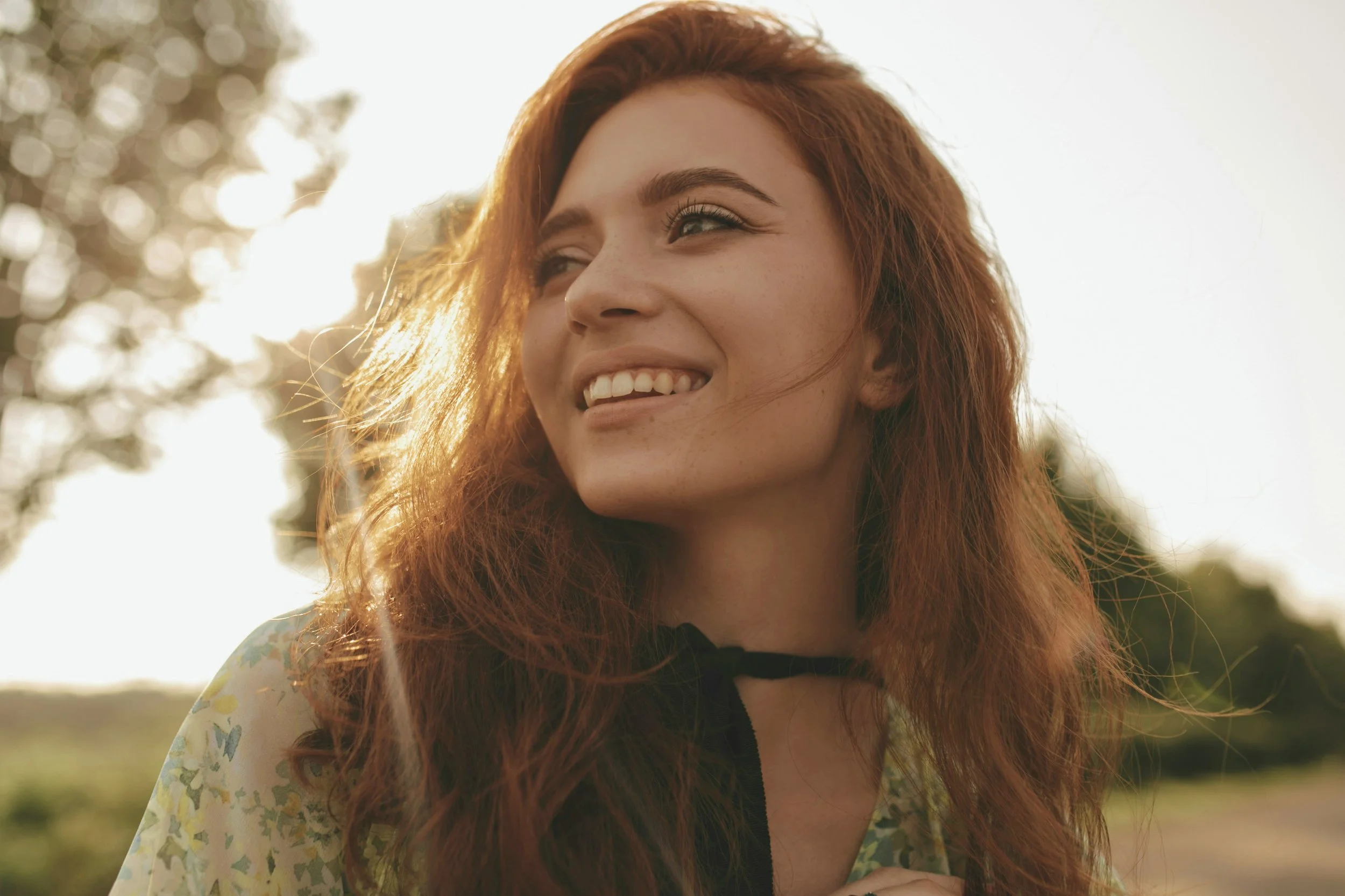 Close-up of a young woman with long red hair, smiling outdoors in sunlight, with blurred trees in background.