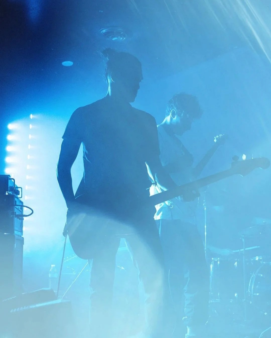 Two musicians perform on stage in blue lighting; one plays an electric guitar, and the other stands with a bass guitar.