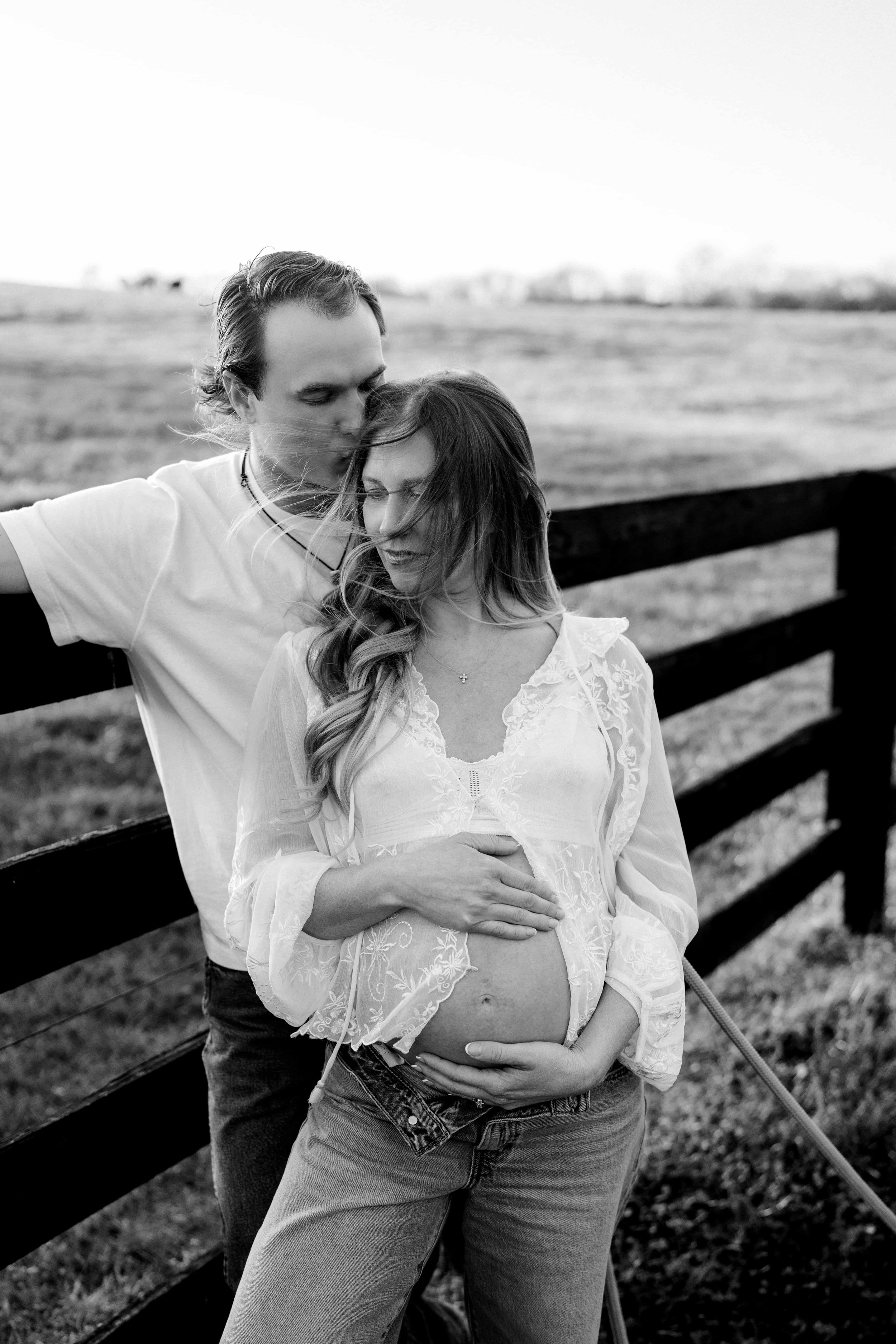 A black and white photo of a pregnant woman with long hair and a patterned blouse holding her belly, standing by a fence. A man with short hair leaning in to kiss her on the forehead with one arm resting on the fence. Outdoors on a grassy field with a blurry landscape in the background.