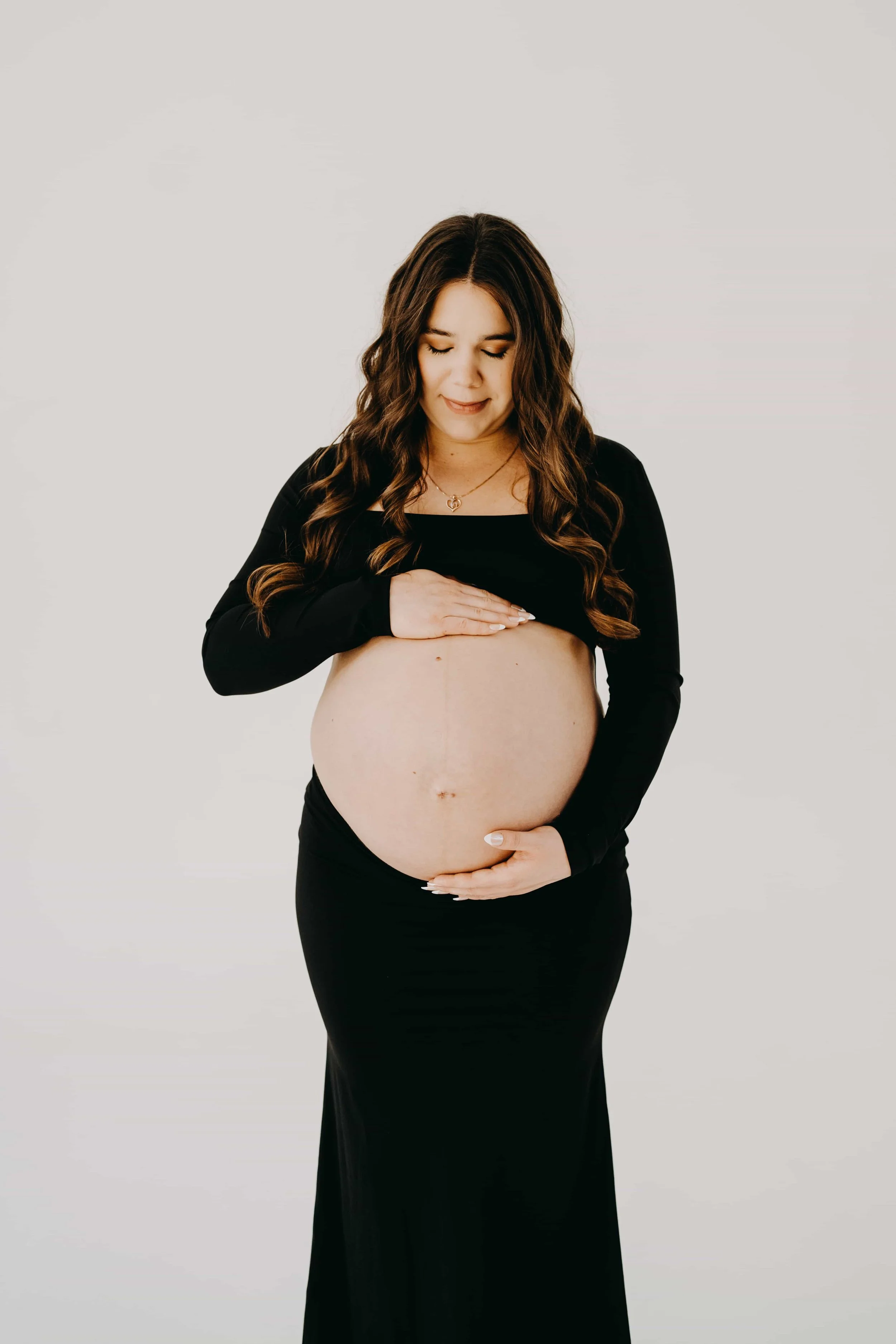 Pregnant woman with long wavy brown hair wearing a black dress and top, gently holding her belly with both hands, standing against a plain white background.