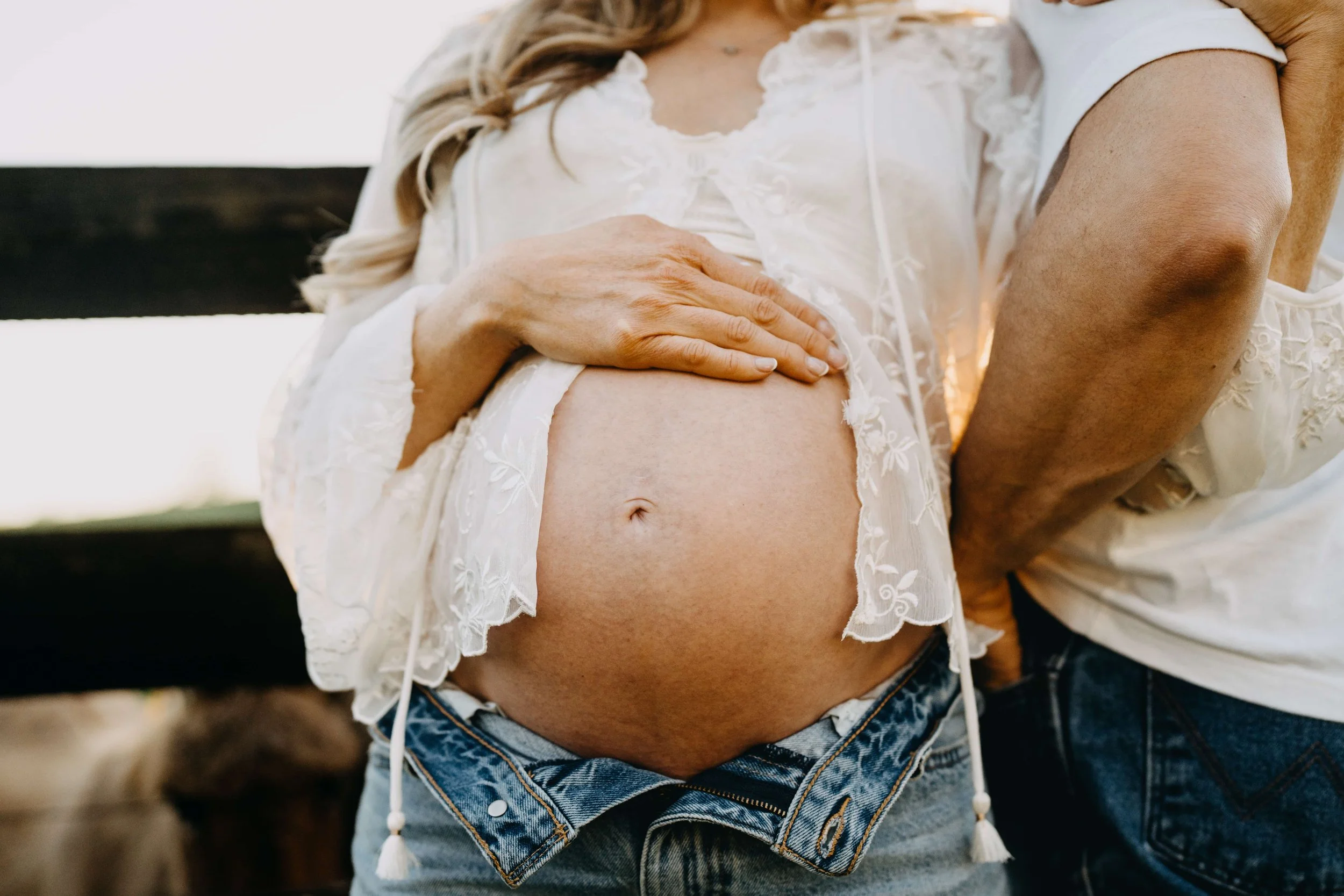 Close-up of pregnant woman's belly being gently touched by another person, both wearing light-colored clothing with lace details, outdoors with blurred background.
