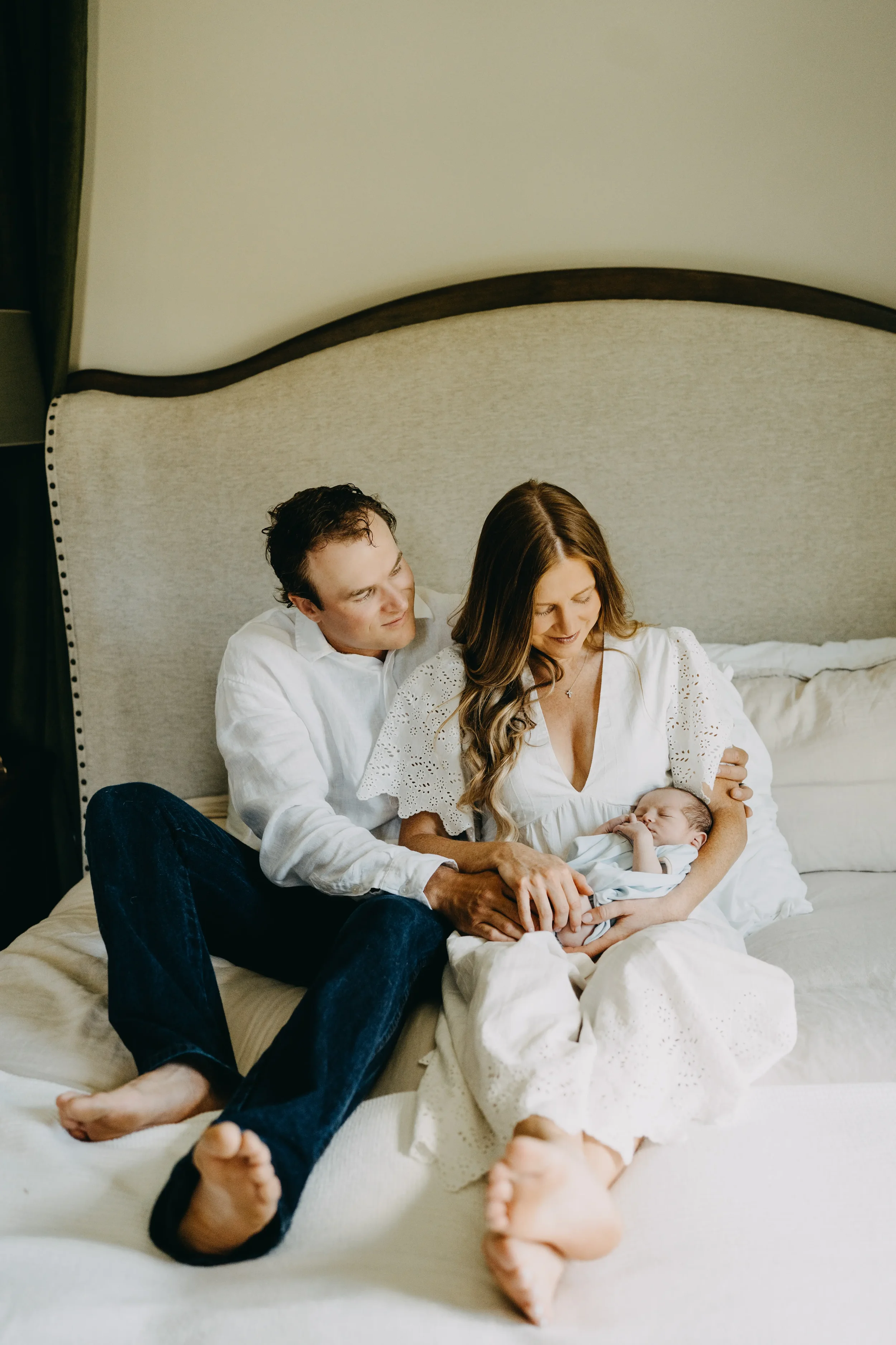 A couple and their baby sitting on a bed with white bedding and a large cream-colored headboard, family enjoying a moment together.