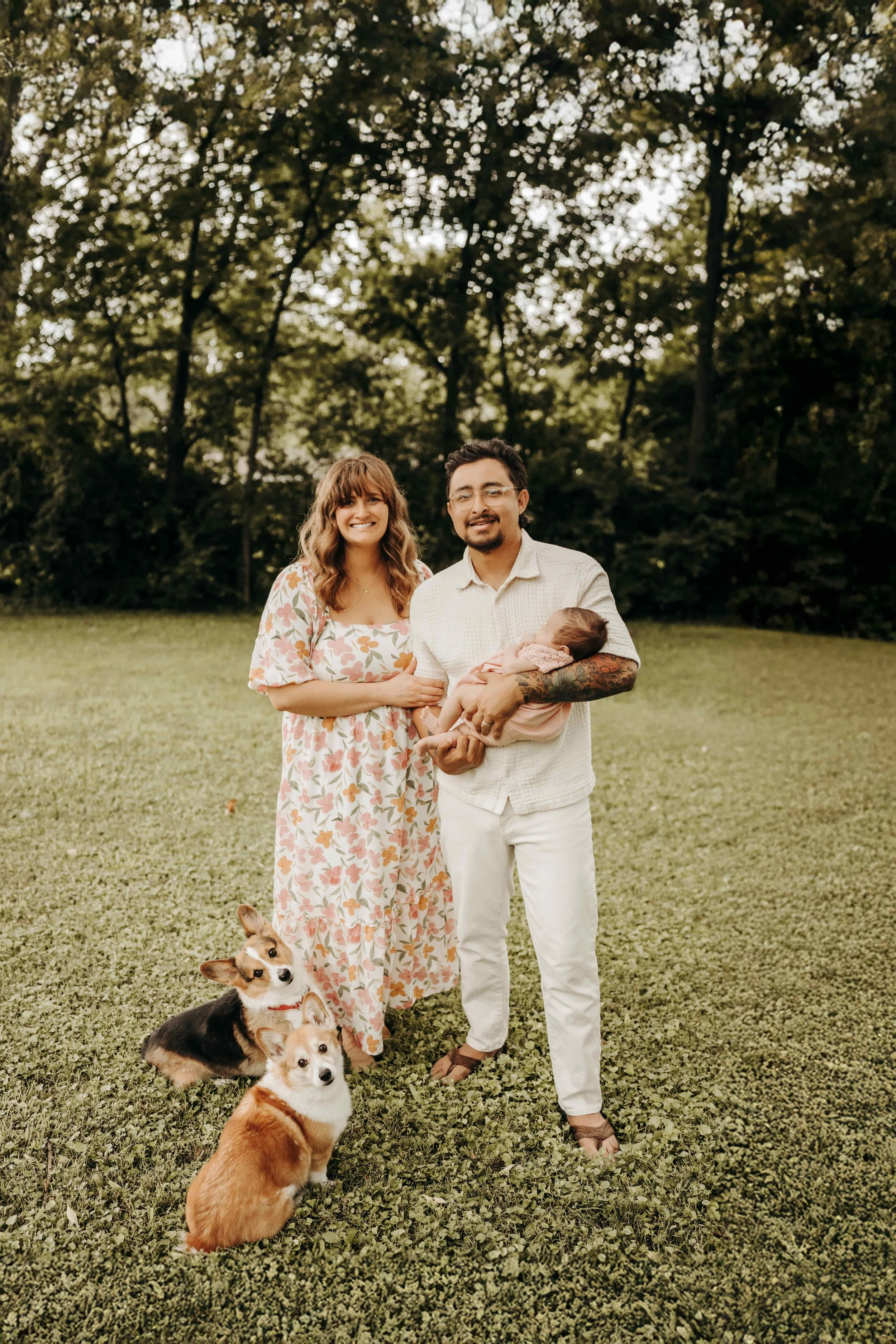 A family of three with two dogs standing on a grassy field with trees in the background. The woman is wearing a floral dress, the man is holding a baby, and the dogs are sitting on the grass.