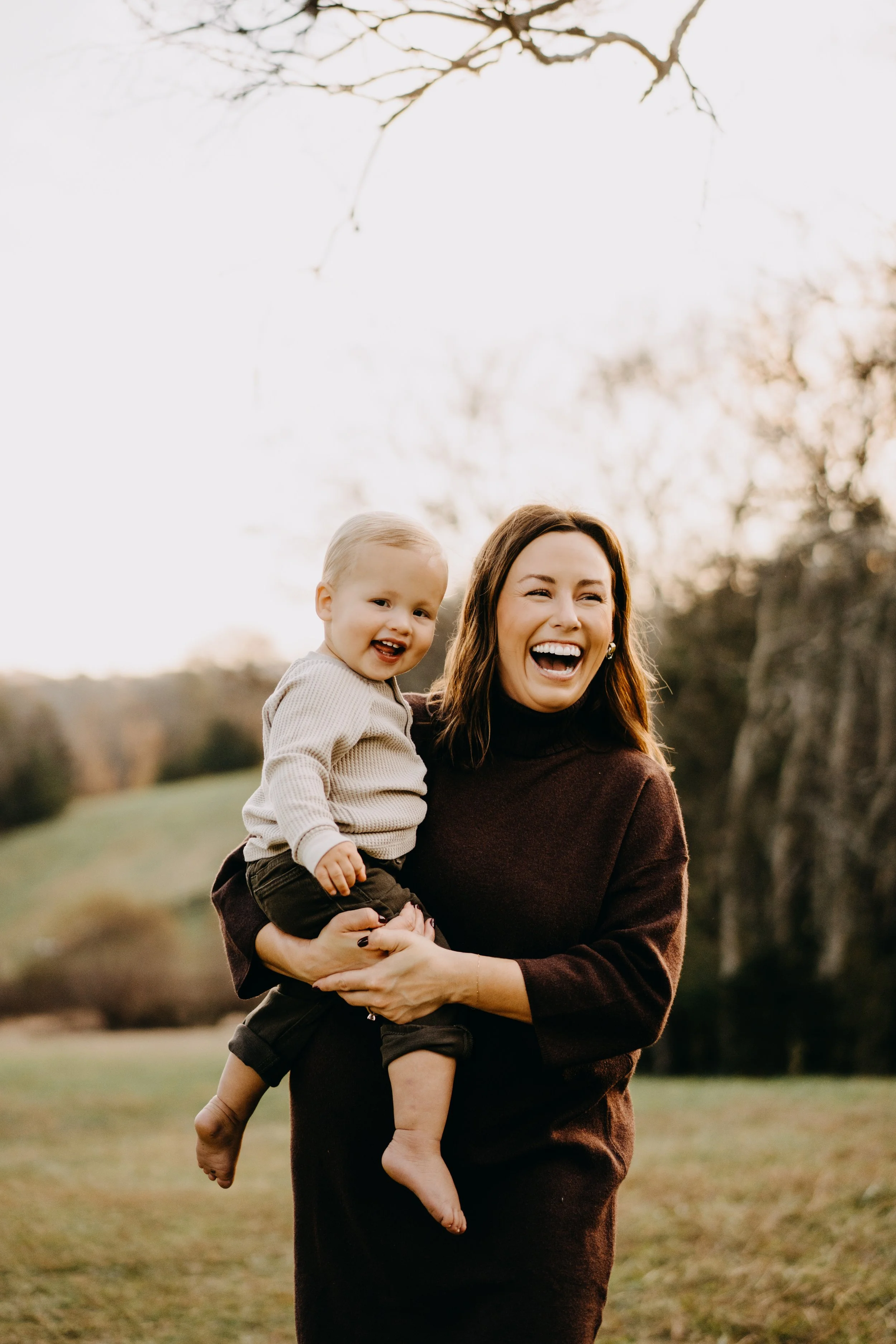 A woman holding a smiling toddler outdoors during sunset, with trees in the background.
