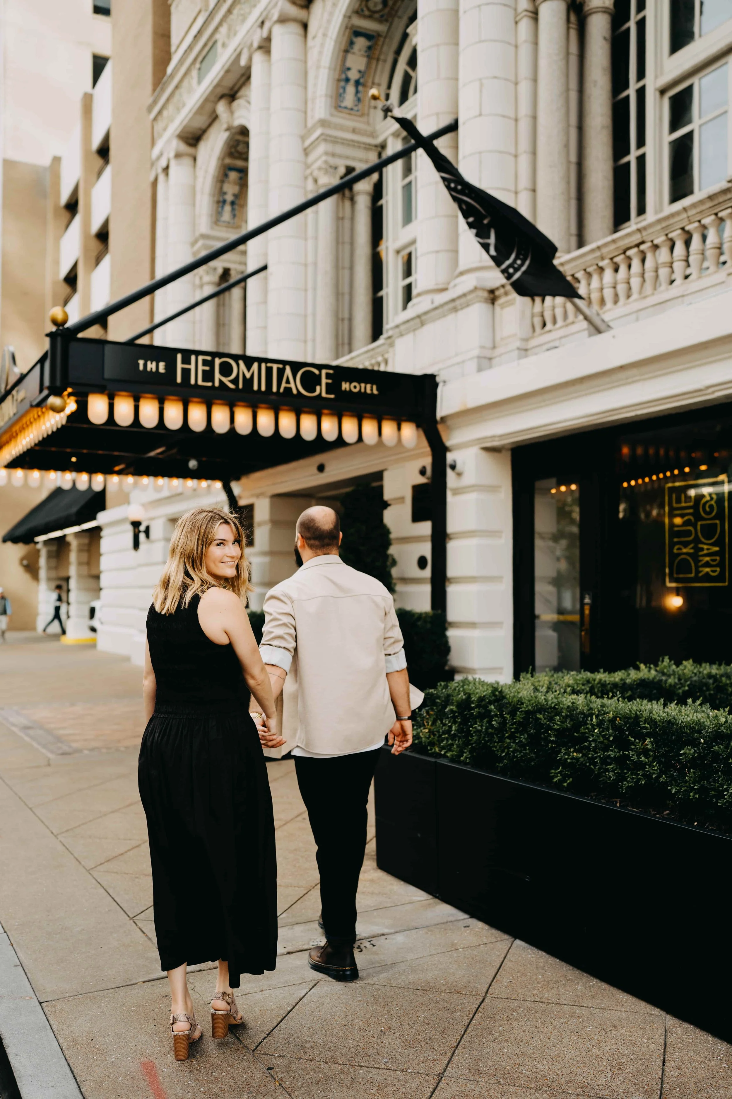 A couple walks hand-in-hand past the Hermitage Hotel's entrance with marquee lights during the evening, with the woman smiling at the camera.