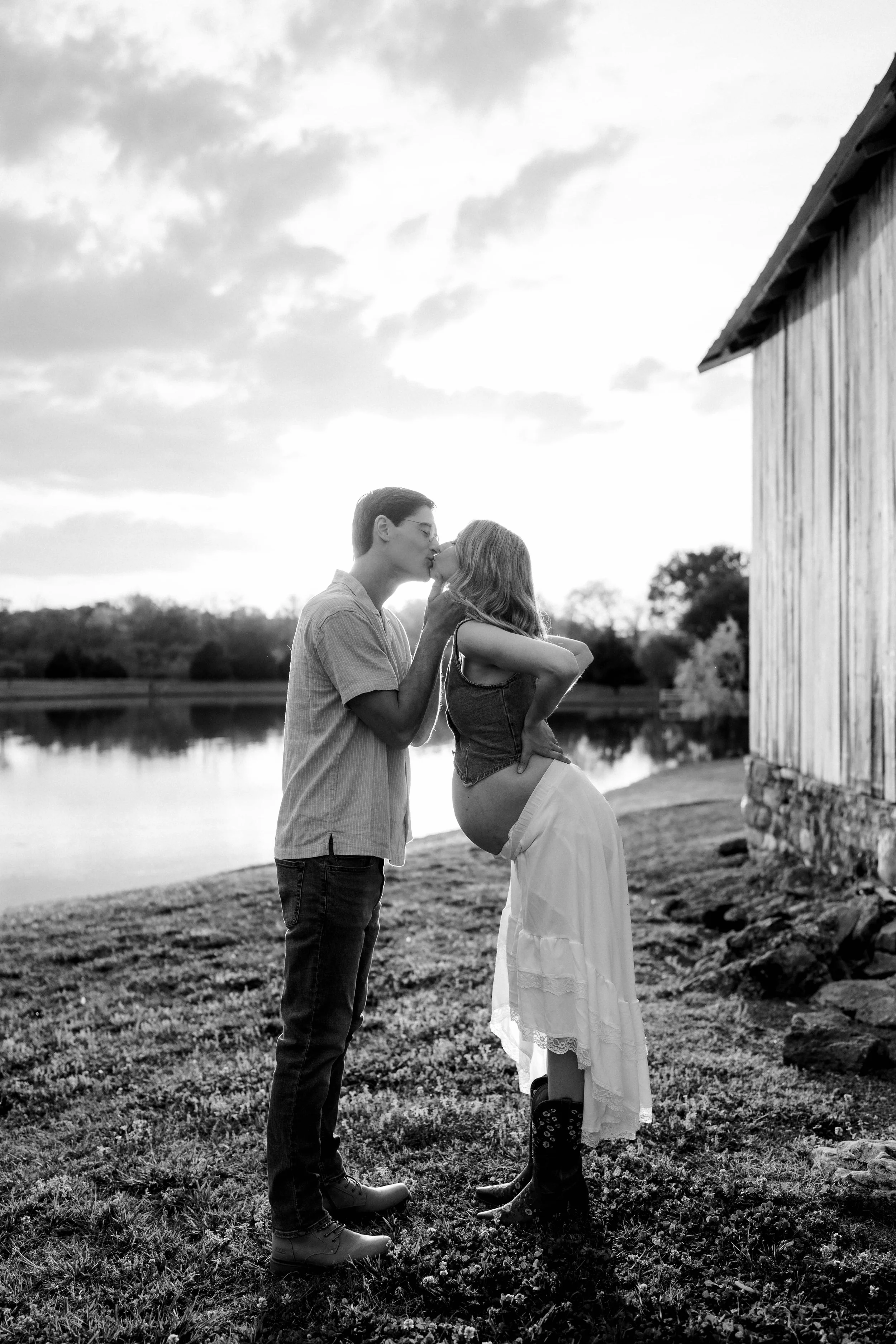A black-and-white photo of a man and a woman sharing a kiss outdoors near a pond. The man is wearing glasses, a short-sleeved shirt, jeans, and boots. The woman, who appears pregnant, is wearing a sleeveless top, a long skirt, and boots. They are standing close to a rustic building with a wooden exterior and stone foundation, with trees and a cloudy sky in the background.