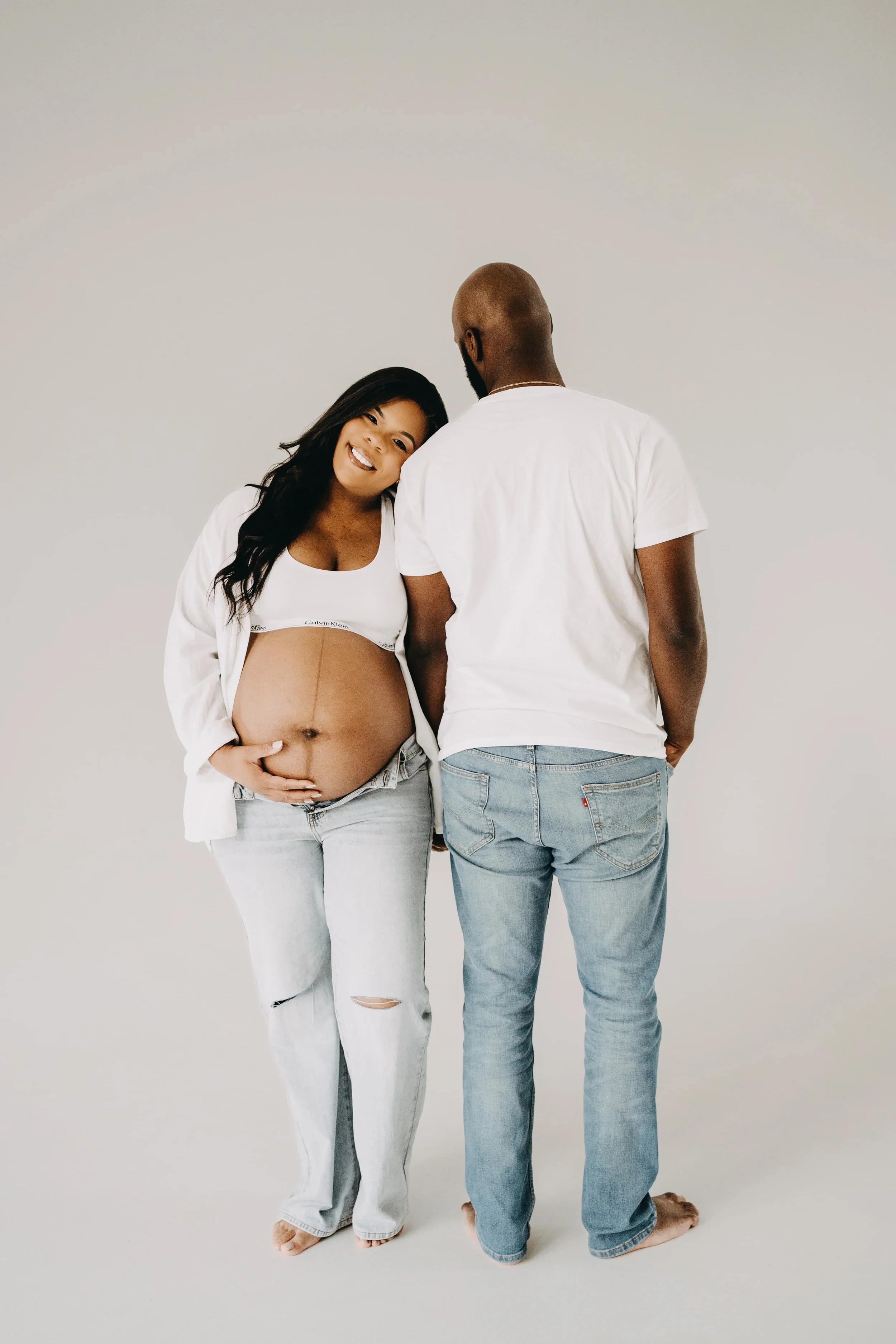 A pregnant woman with long dark hair, smiling and touching her belly, standing next to a man with a shaved head and beard, both dressed in white shirts and light jeans, facing each other against a plain light background.