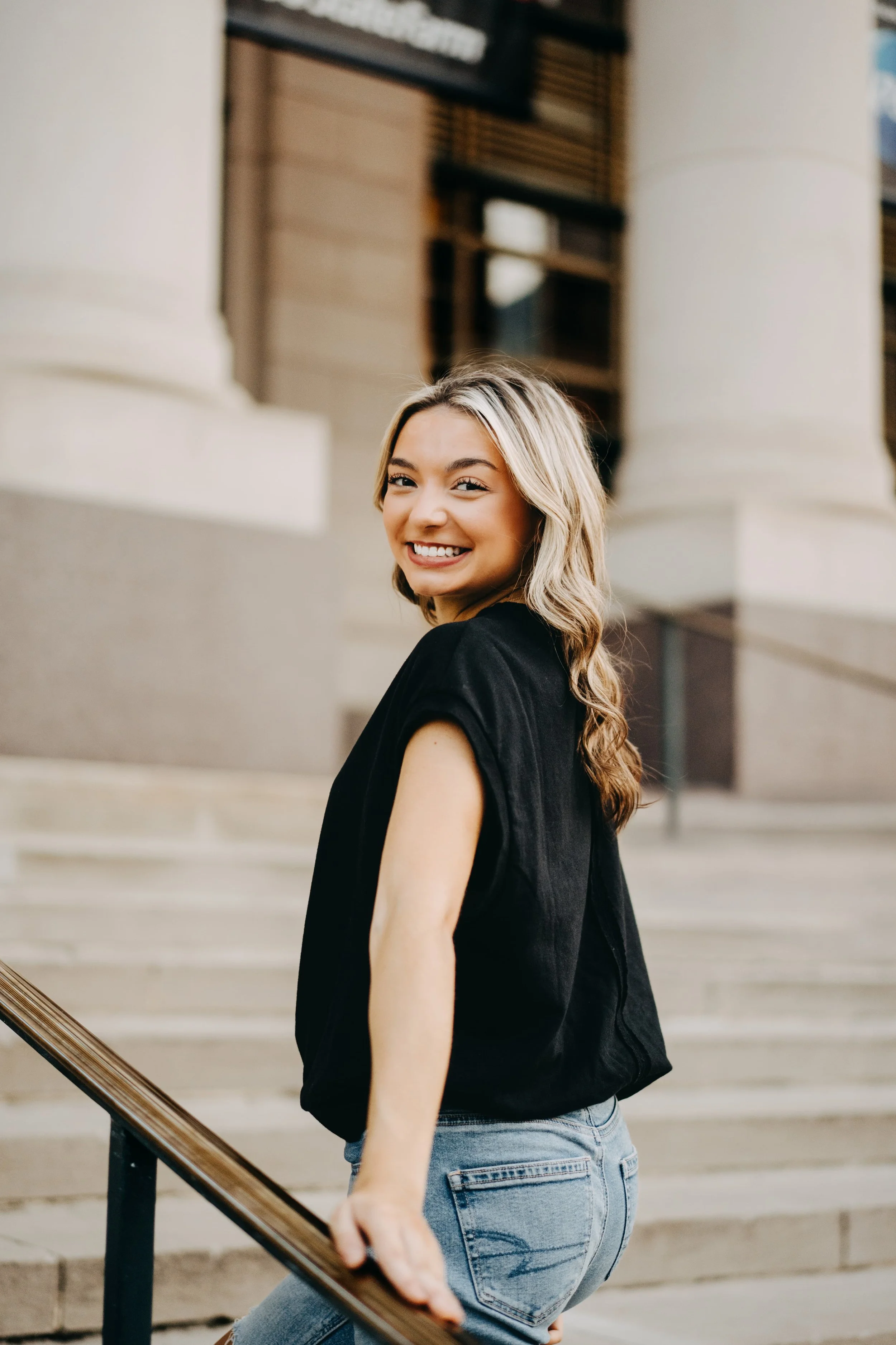 A young woman smiling and looking over her shoulder as she holds the railing on outdoor stairs of a classical building.