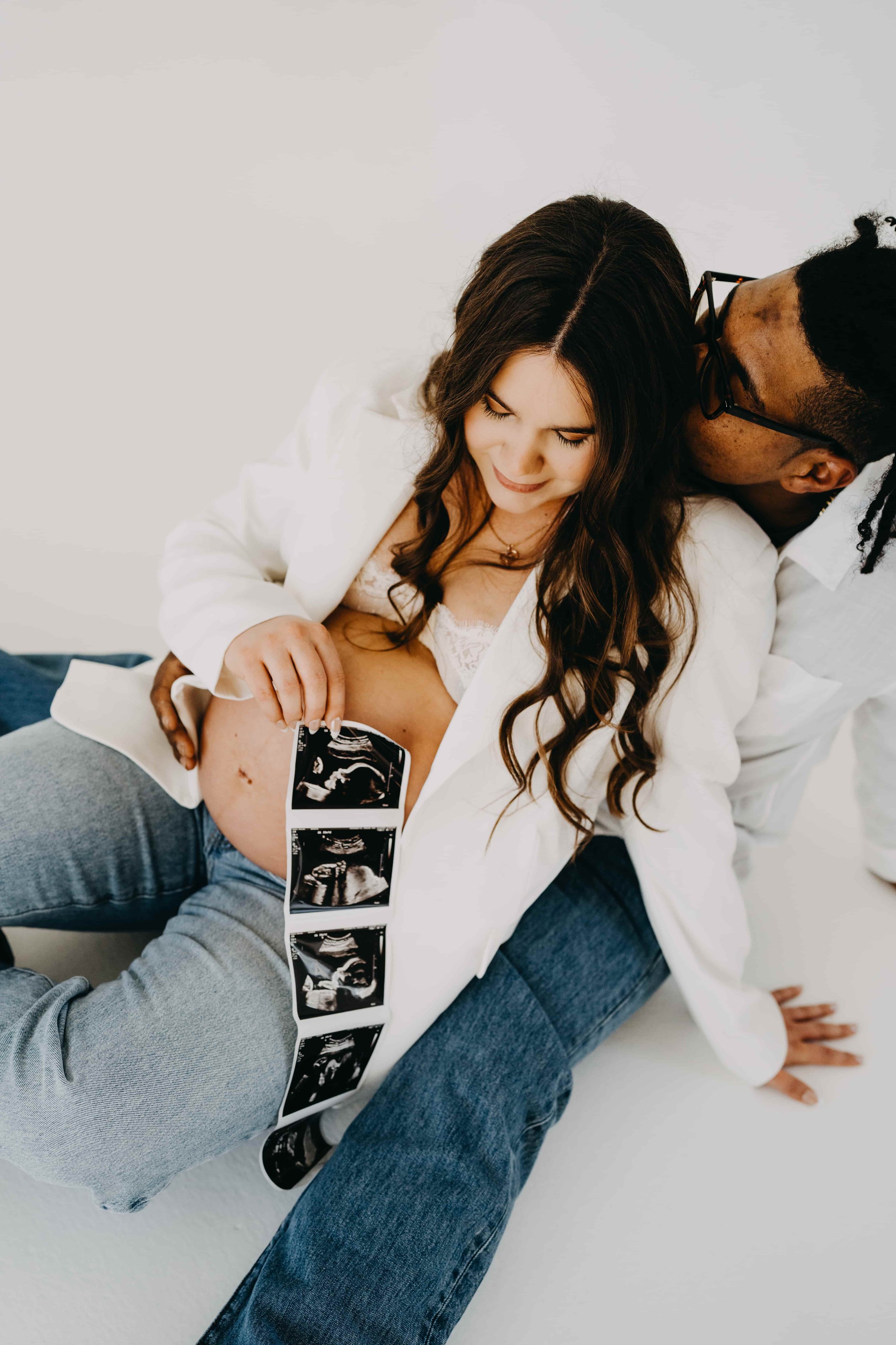 A pregnant woman with long brown hair, smiling, holding an ultrasound scan, with her partner kissing her on the head, sitting on a white surface.