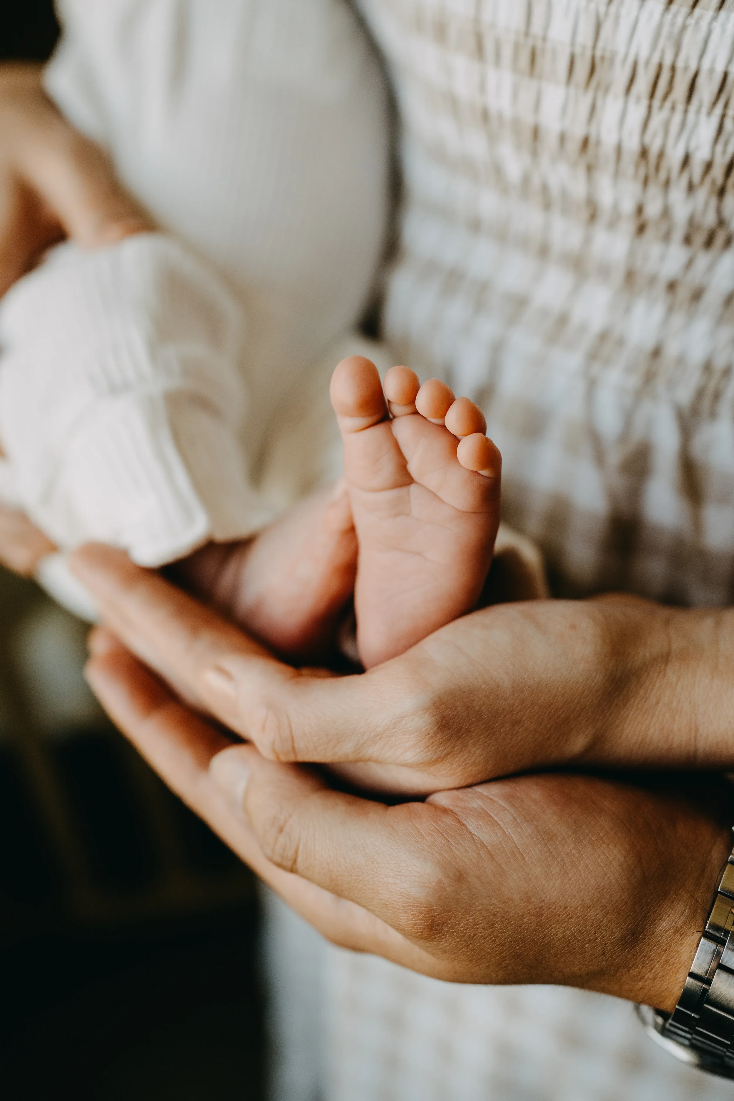 A close-up of an adult's hand gently cradling the tiny hand of a baby, with the baby wearing a white, striped long-sleeve outfit. The adult's wrist has a silver watch.