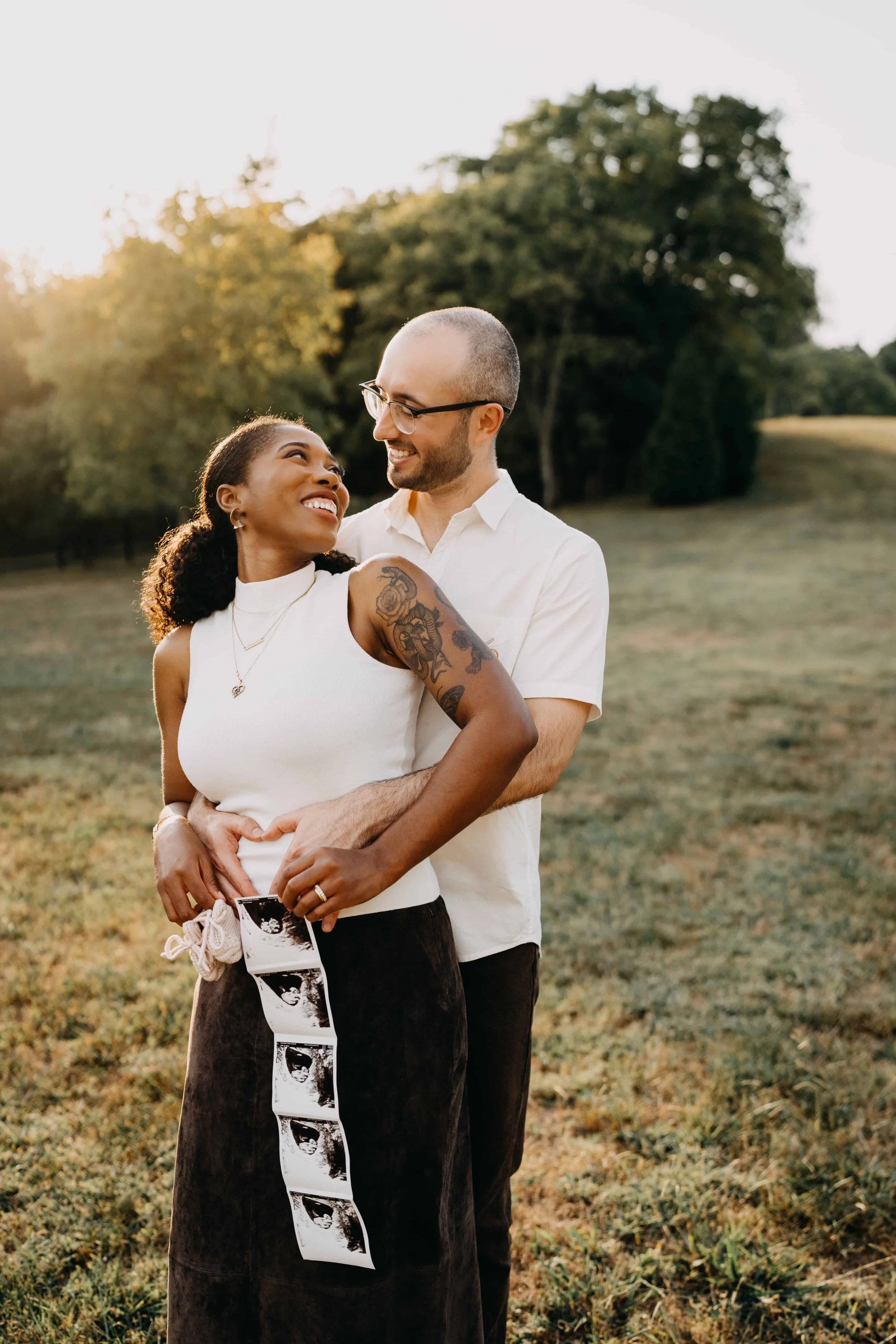 A happy couple stands outdoors in a grassy field during sunset, smiling and looking at each other. The woman is holding ultrasound pictures, and the man is embracing her from behind.
