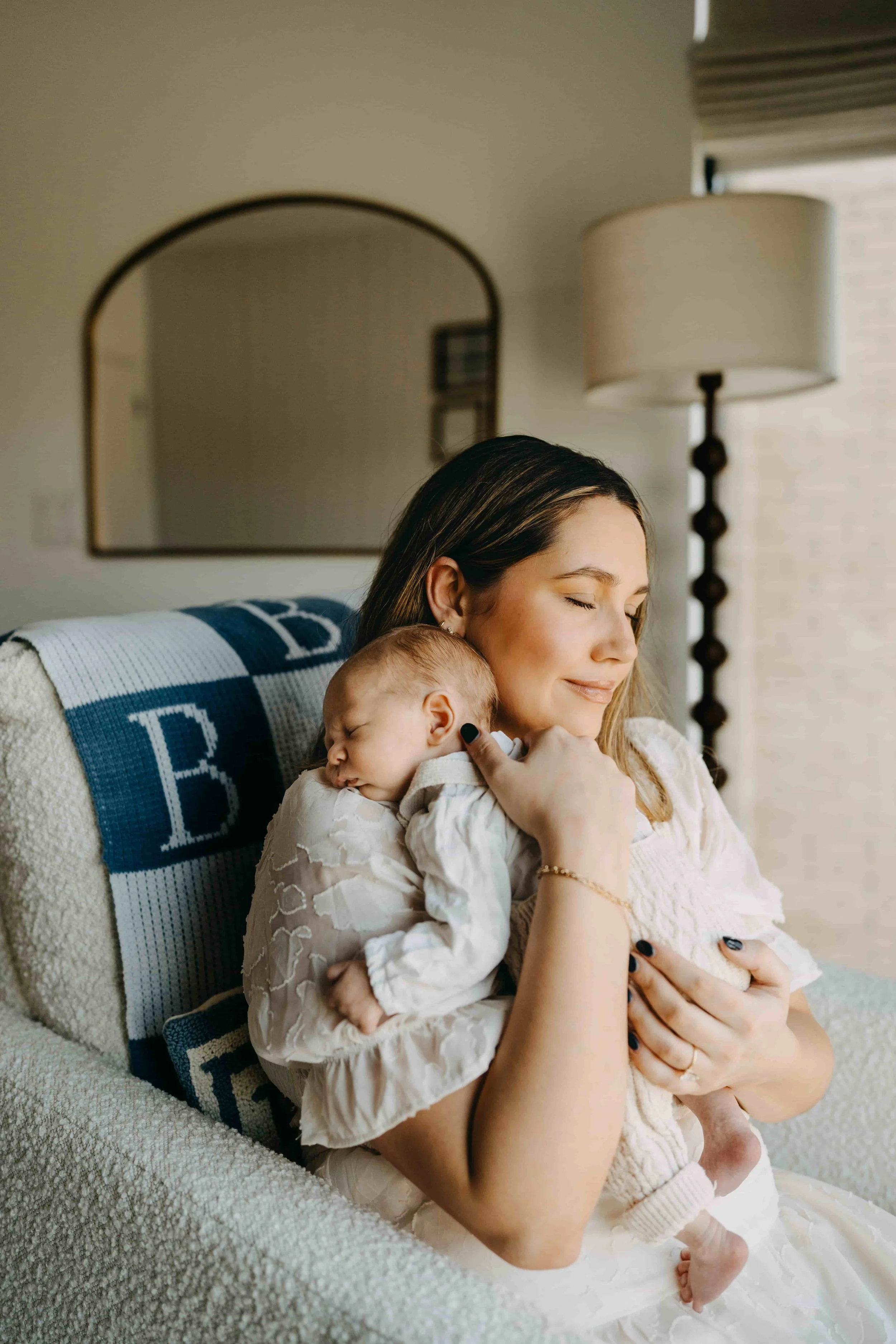 A woman with closed eyes, smiling peacefully, cuddles a sleeping baby on her shoulder while sitting on a beige couch in a cozy home setting.