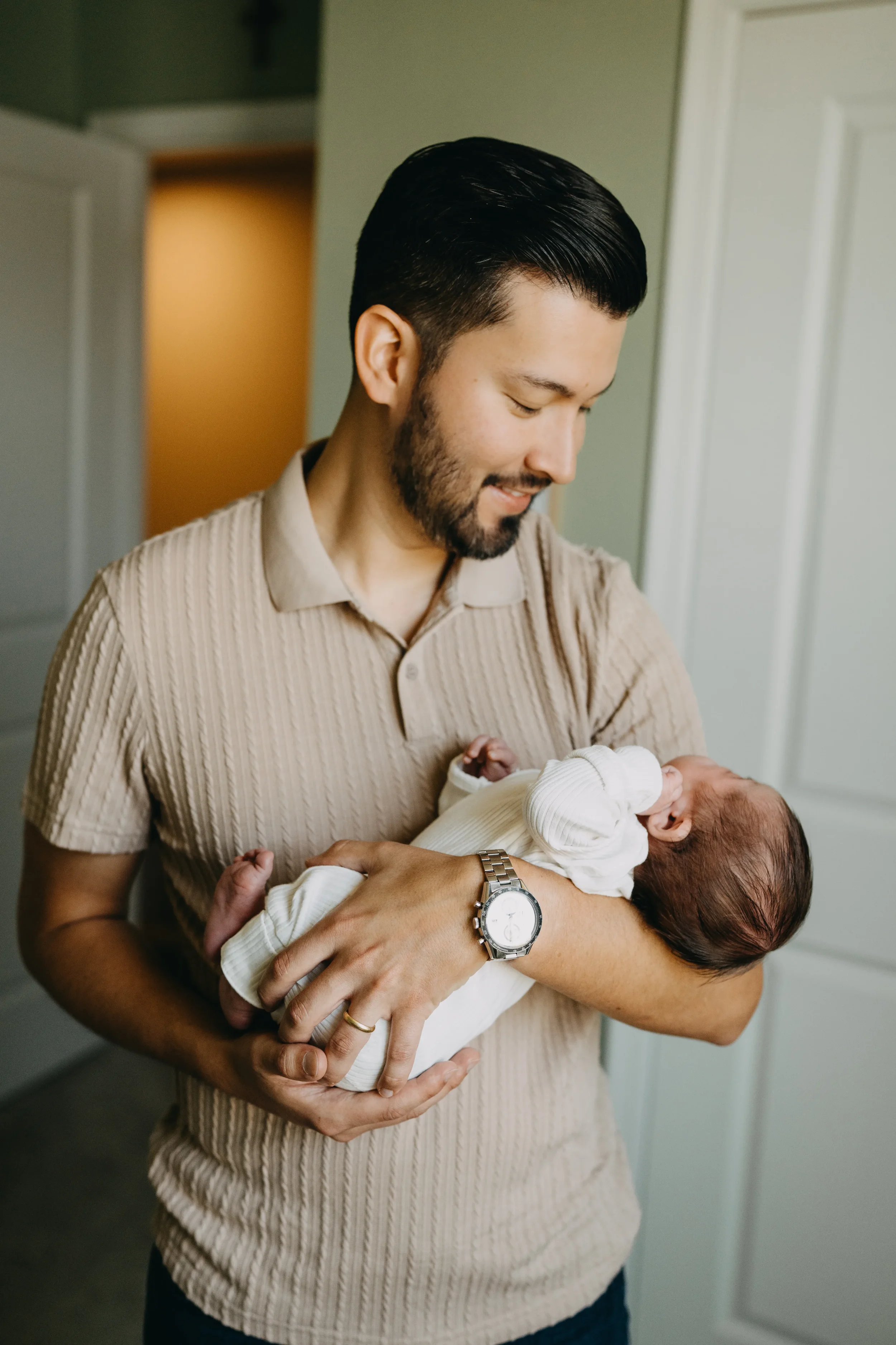 A man with dark hair and a beard holding a newborn baby, smiling and looking down at the baby, indoors in a home setting.