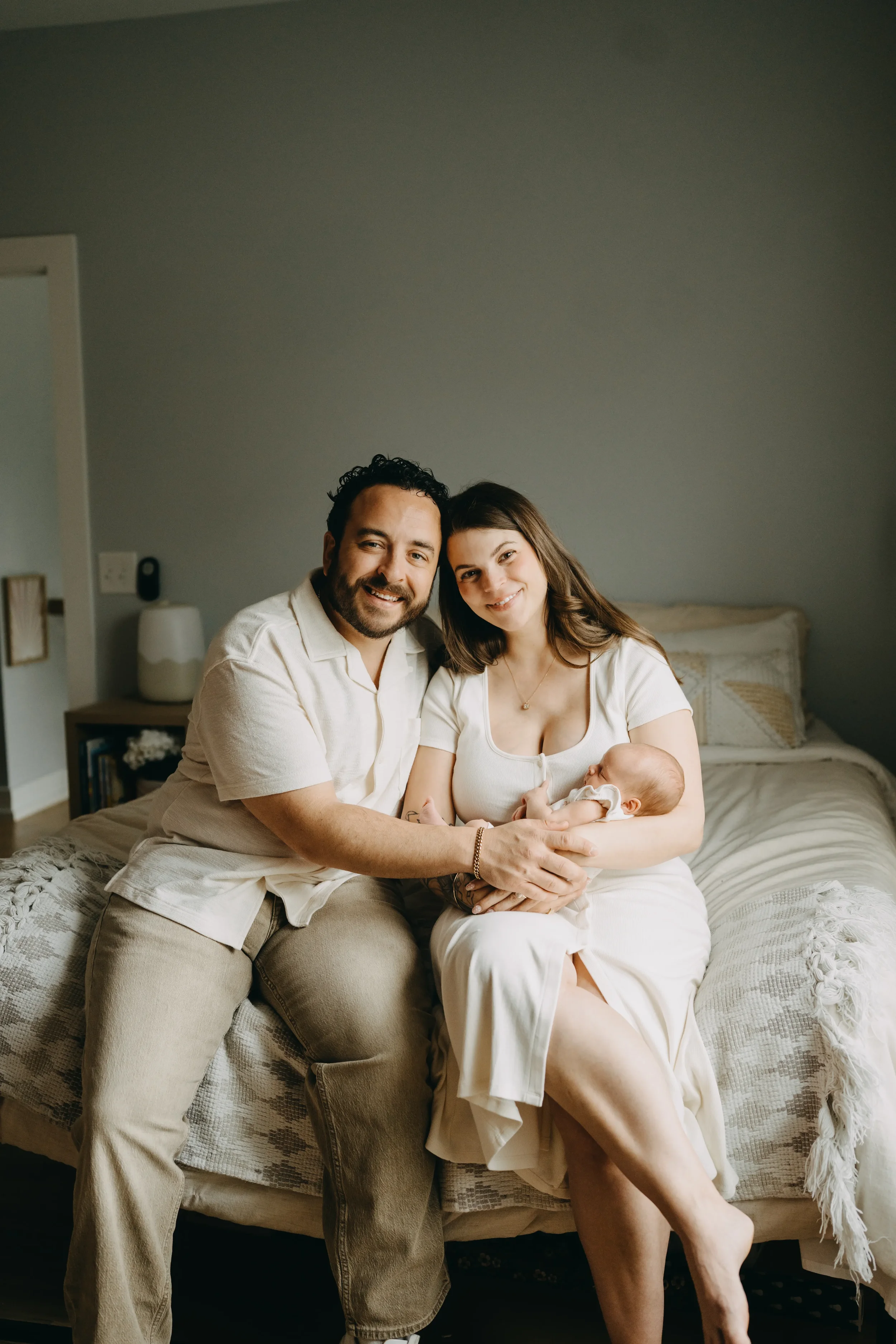 A happy family of three sitting on a bed in a bedroom, holding their newborn baby.