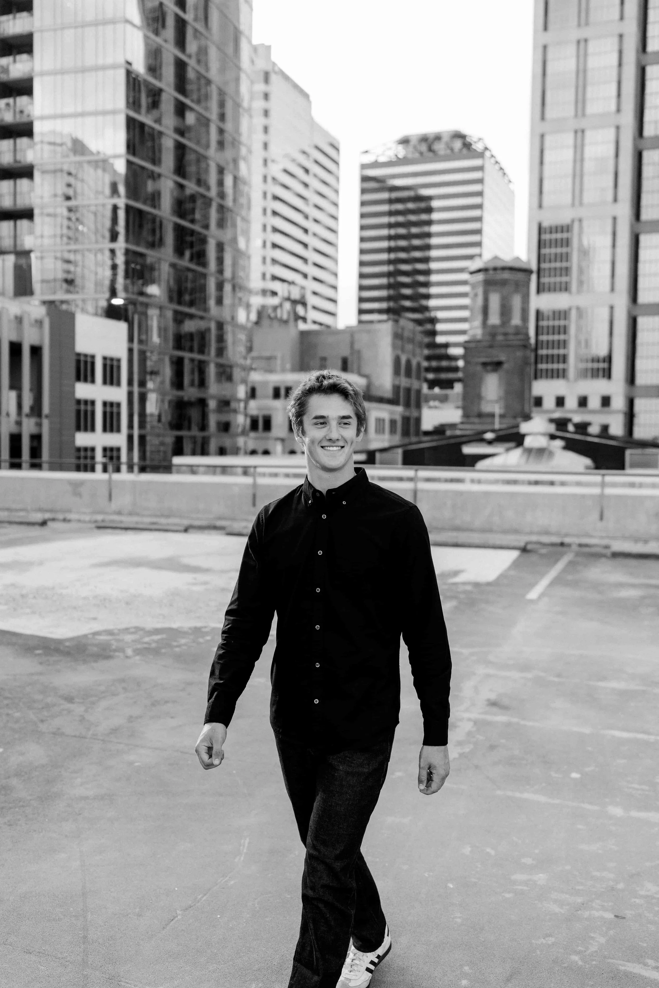A young man in dark clothing, smiling and walking in an urban rooftop parking lot with skyscrapers in the background.
