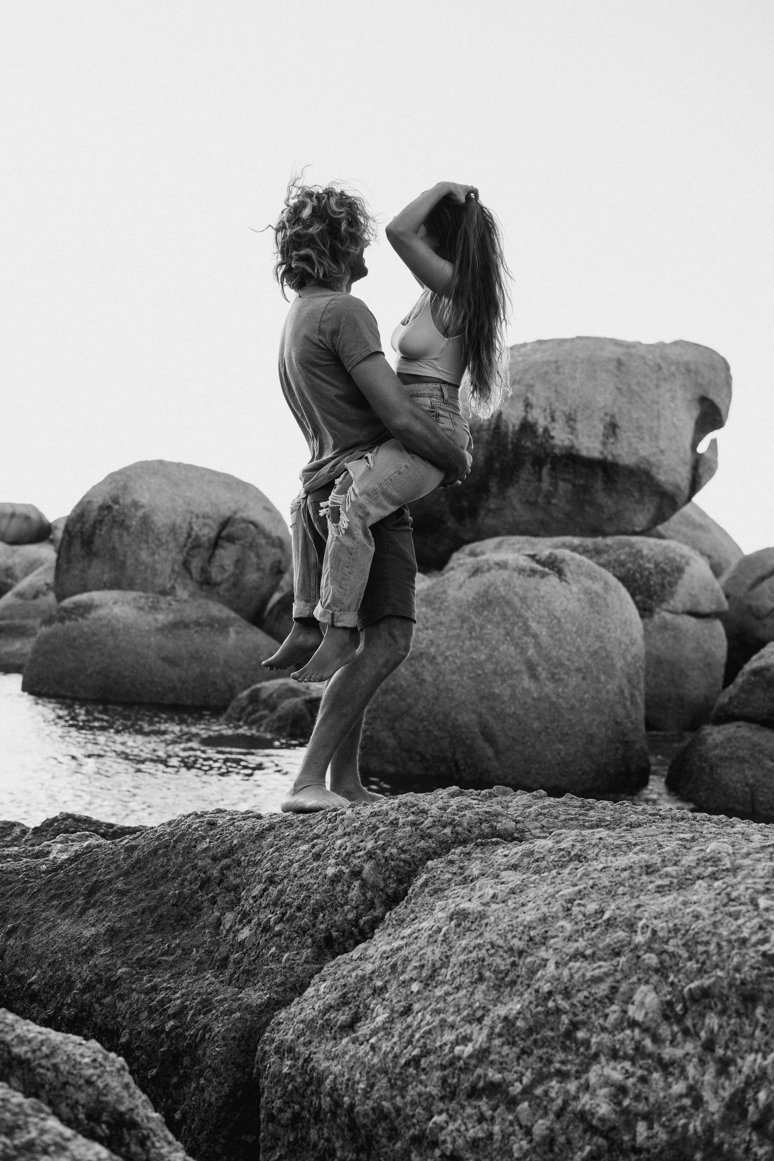A black and white photograph of a man holding a woman in his arms on a rocky shoreline with large boulders in the background.