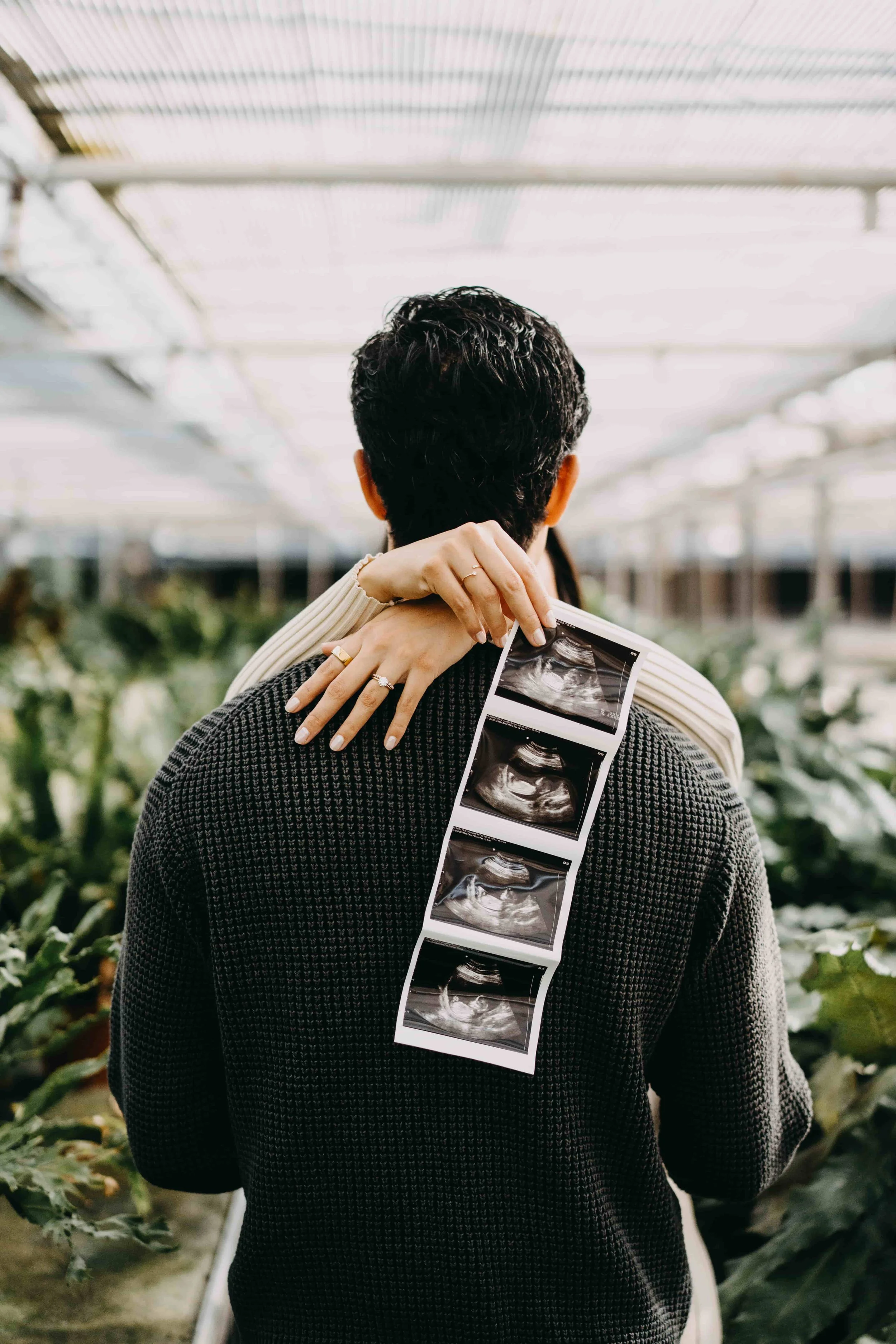 Couple in a greenhouse, embracing, holding ultrasound photos displaying pregnancy.
