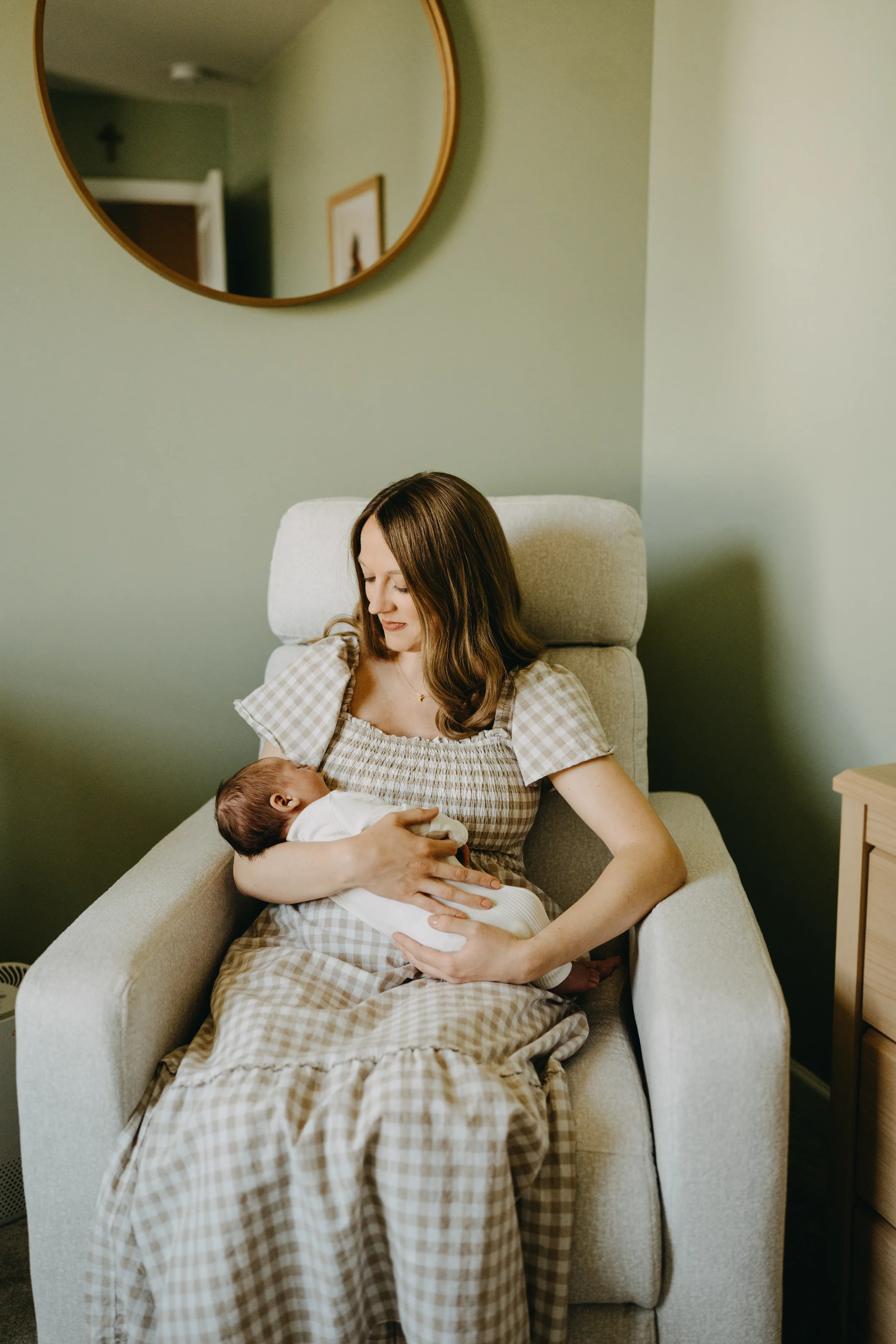 A woman sitting in a beige armchair holding a newborn baby in her arms. She is smiling and looking down at the baby, who is dressed in white clothing. The woman is wearing a checkered dress, and the background features a green wall with a round mirro