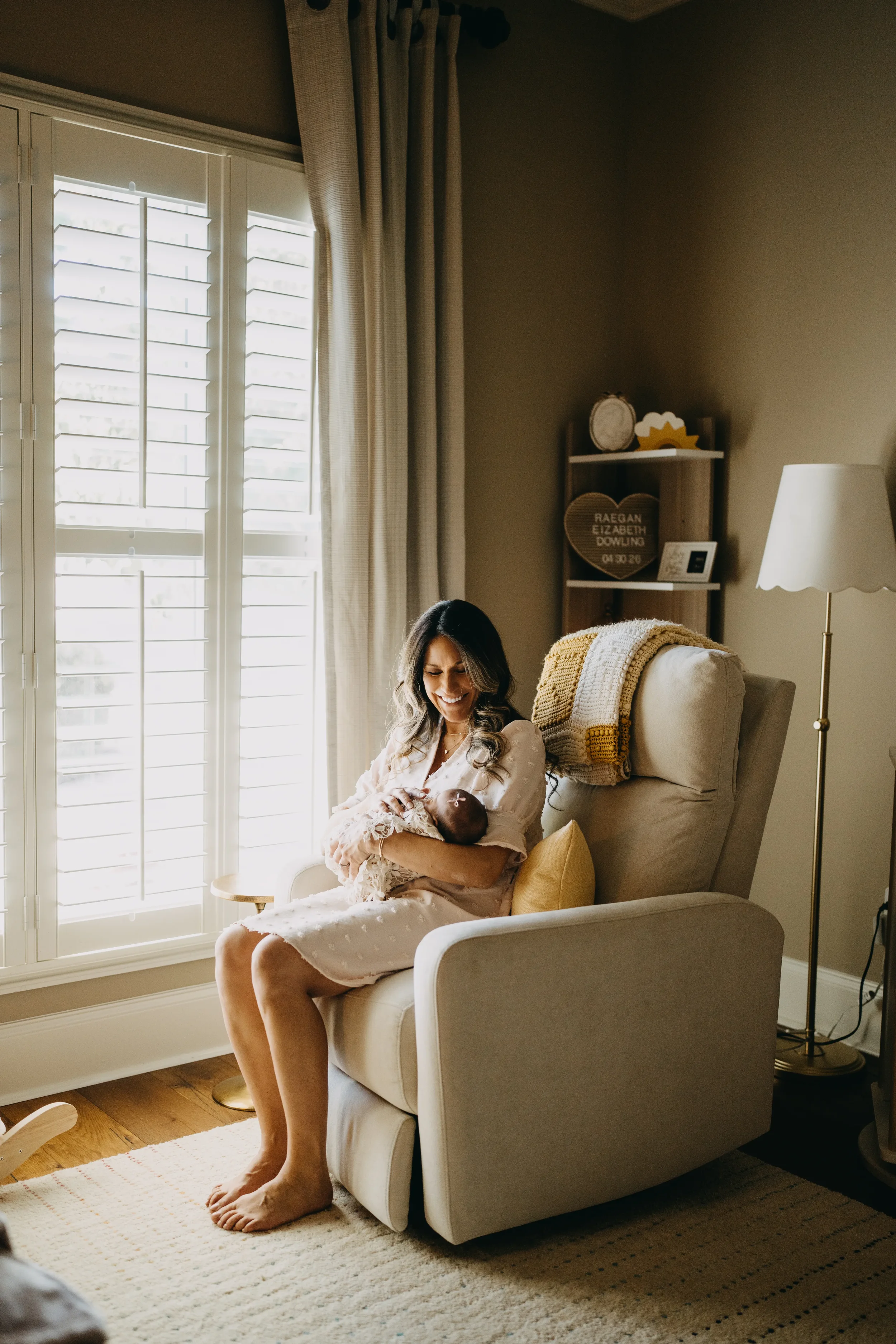 A woman sitting in a light-colored armchair by a window with white shutters, holding a baby wrapped in a blanket. She is smiling and wearing a light pink nightgown. There is a yellow pillow behind her back, and a cozy throw blanket is draped over the arm of the chair. A floor lamp with a white shade stands nearby, and a small shelf with decorative items and framed photo is visible in the background.