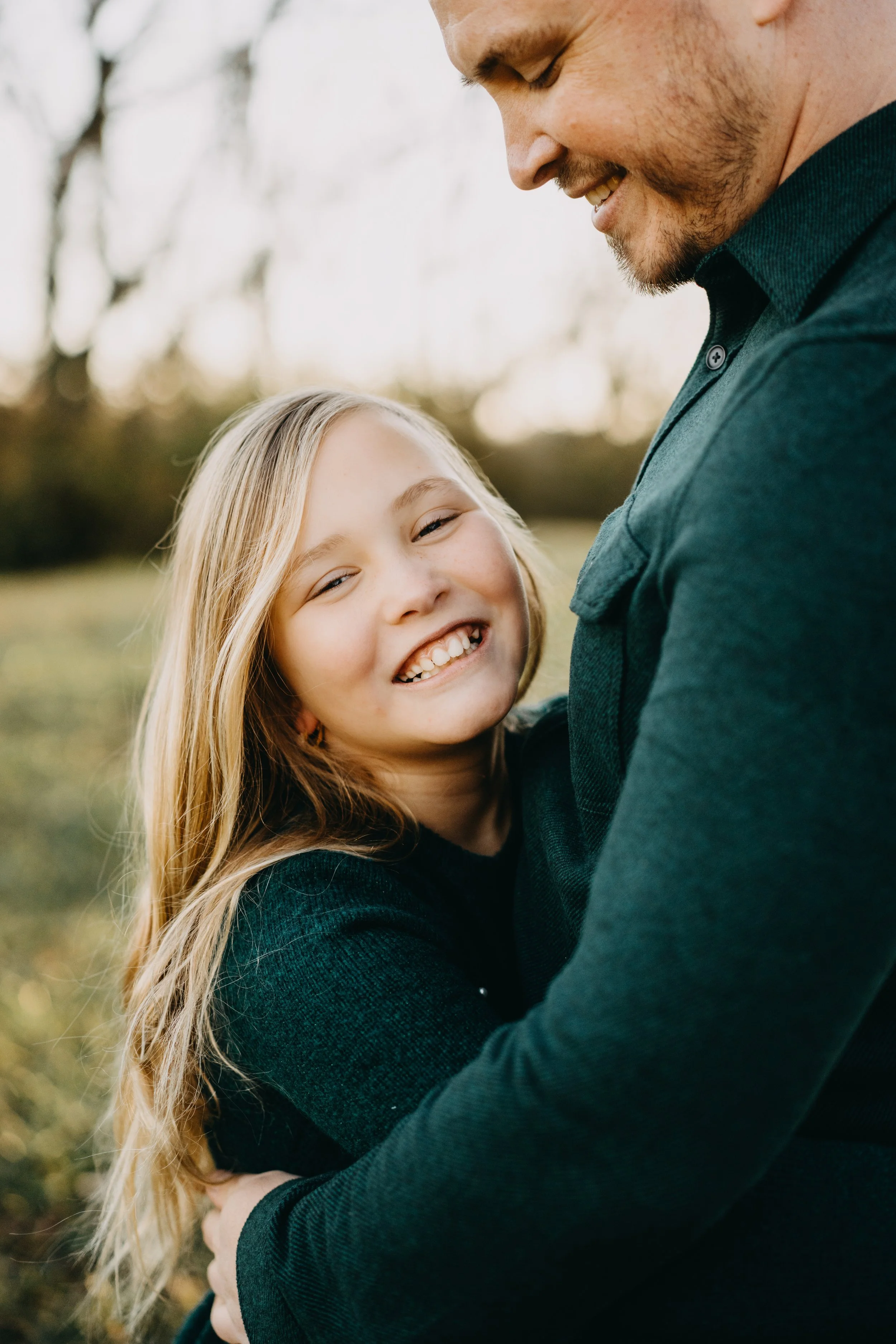 A young girl with long blonde hair smiling and hugging a man outdoors, with trees in the background.