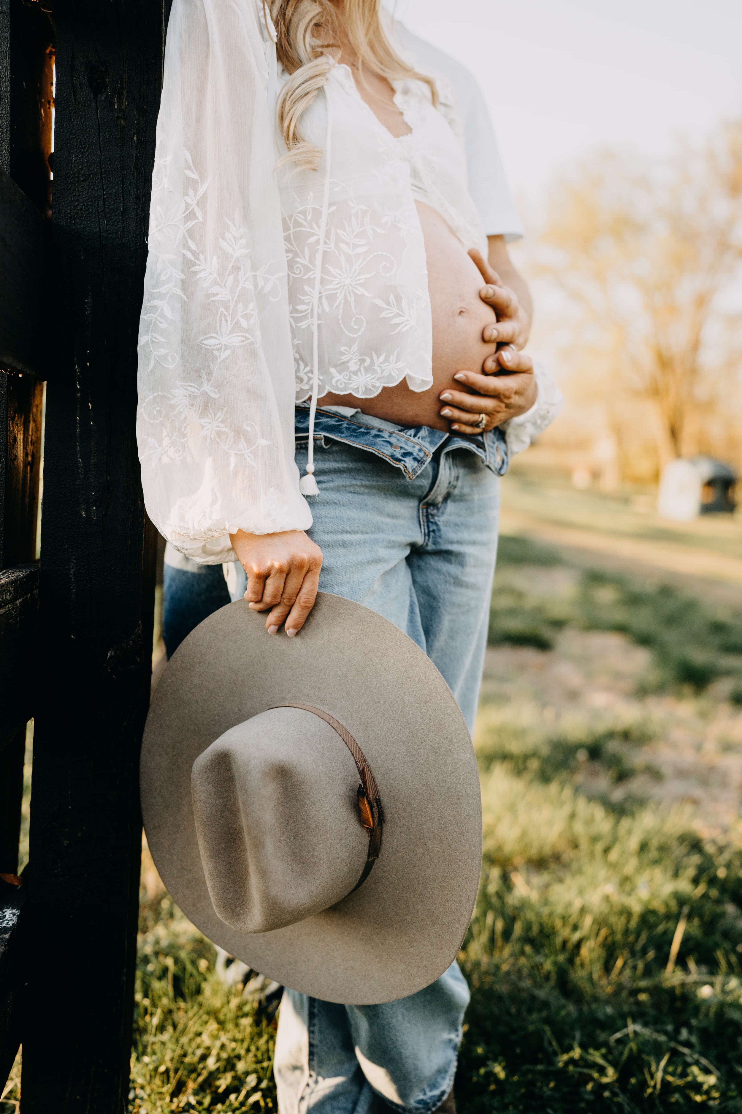 A pregnant woman is leaning against a black fence, holding a wide-brimmed hat in her right hand. Her left hand rests on her belly, with another hand supporting her from underneath. She is wearing a sheer white embroidered top and blue jeans. The background features a natural outdoor setting with trees and sunlight.