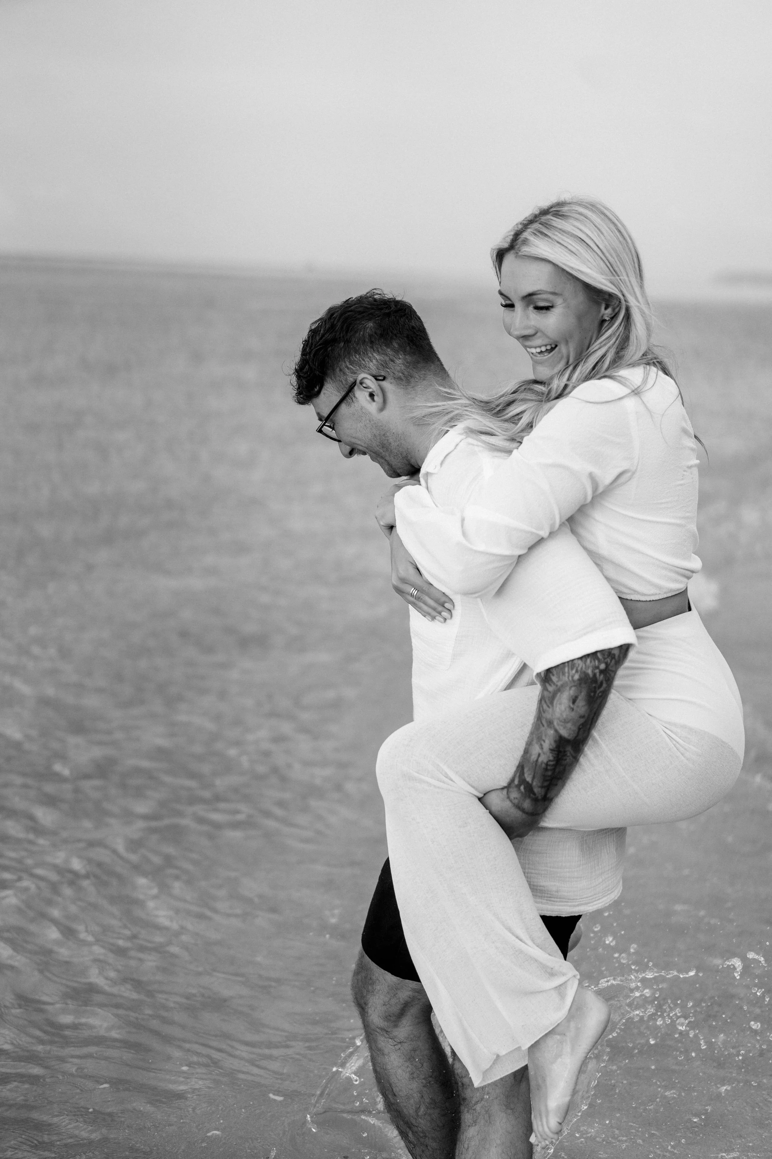 A smiling woman with long blonde hair piggybacking on a man with glasses and tattoos, walking through shallow water at the beach.