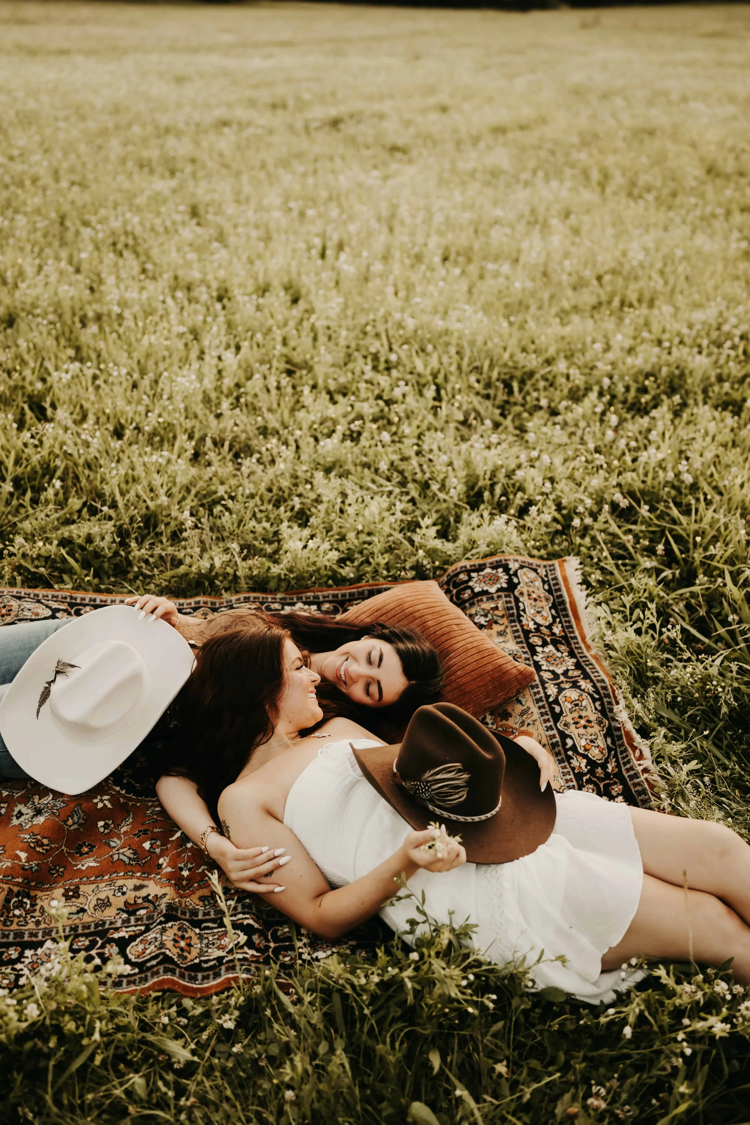 Two women lying on a patterned blanket in a field of greenery, smiling and holding hats.